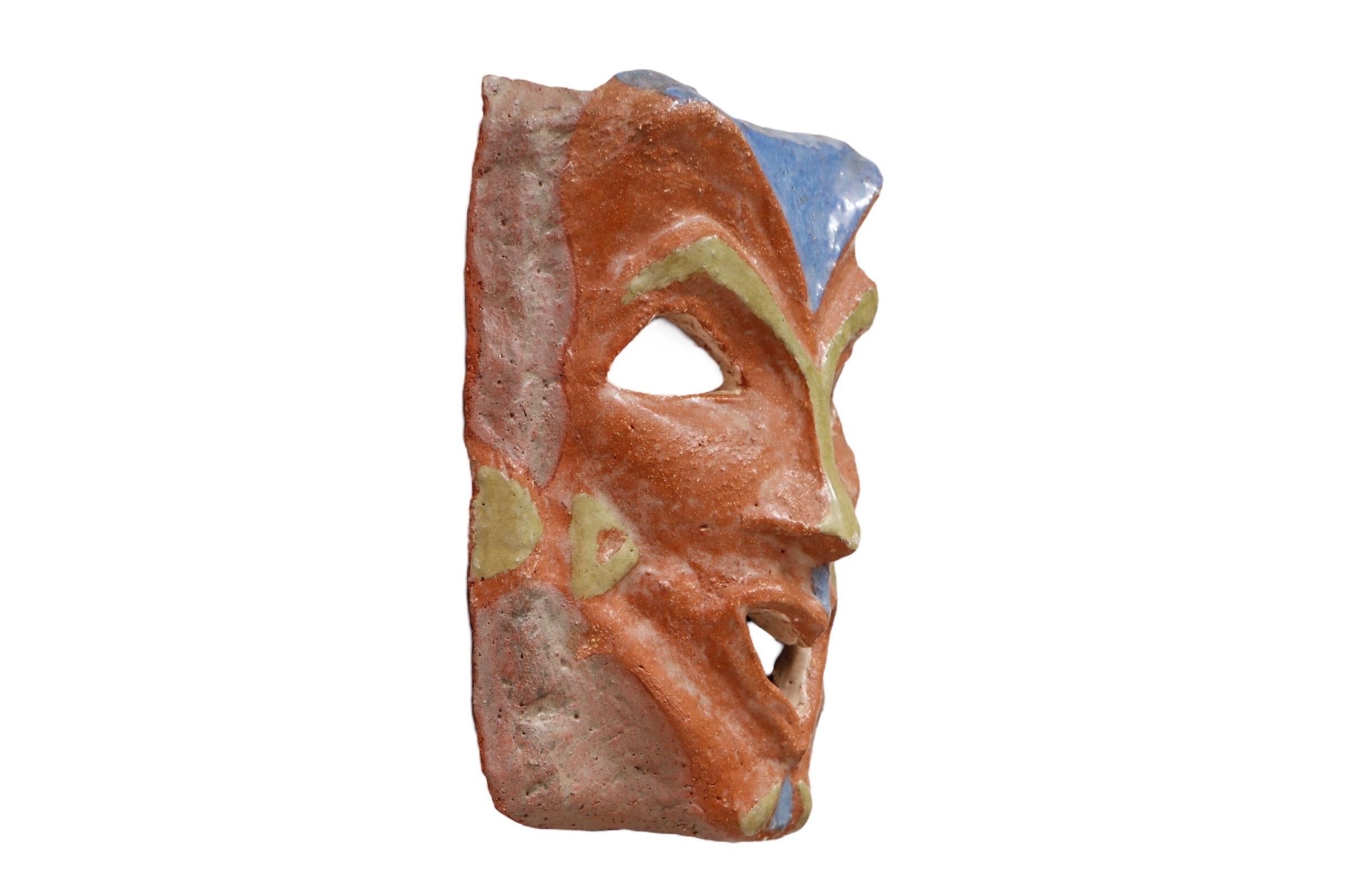 A handmade terracotta pottery art ‘Greek Theatre’ style mask. Angular features are decorated with cerulean blue and olive-green highlights. Arched eyebrows give the look of cat ears, and the mouth is a delicate heart shape. Signed in the back by the