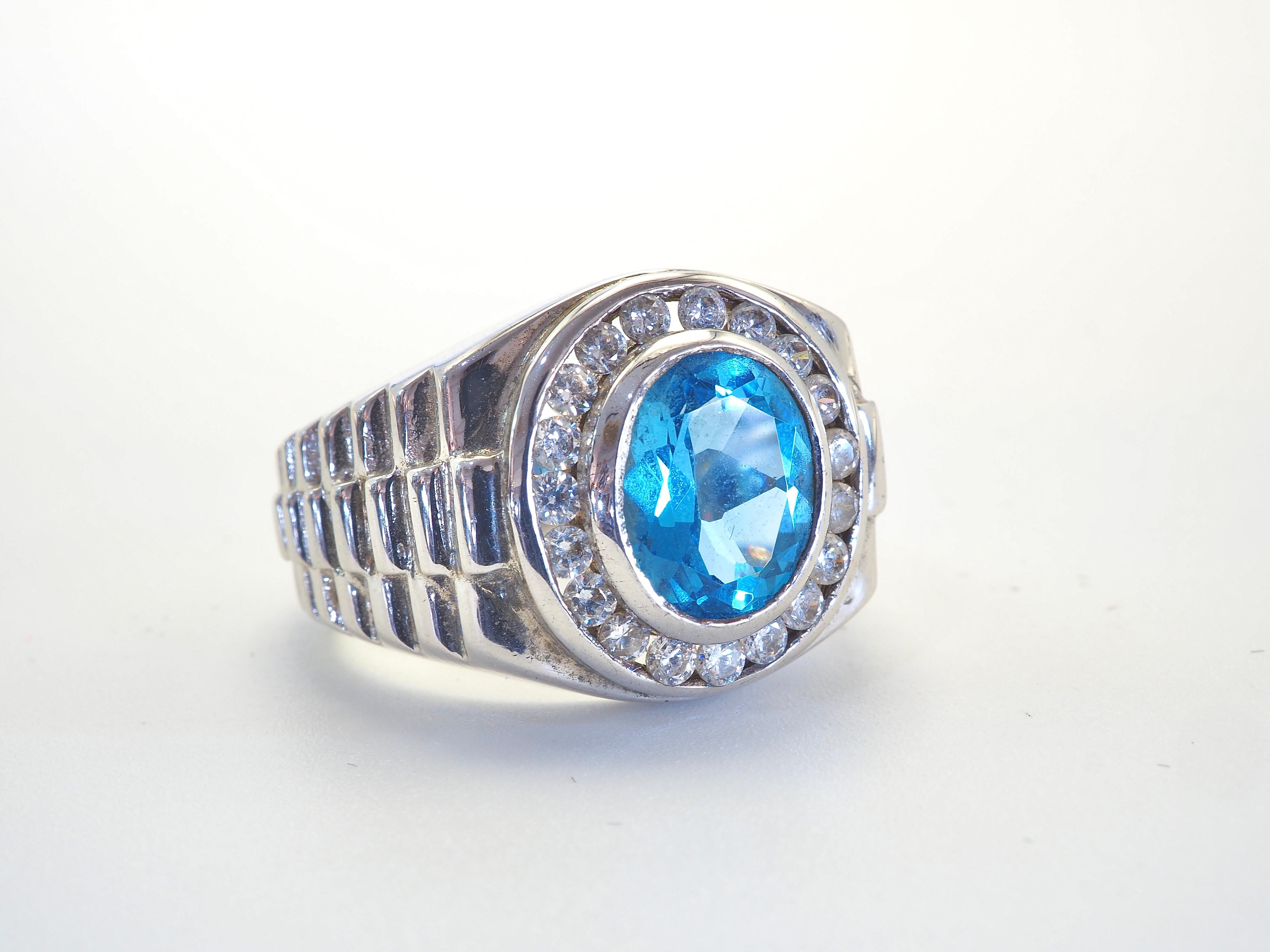 Bague pour homme de belle taille, de type montre-bracelet, dont la pierre principale est une topaze bleue fortement saturée. Les pierres blanches qui l'entourent sont des quartz. 

Nous pouvons redimensionner pour vous pour 15 $