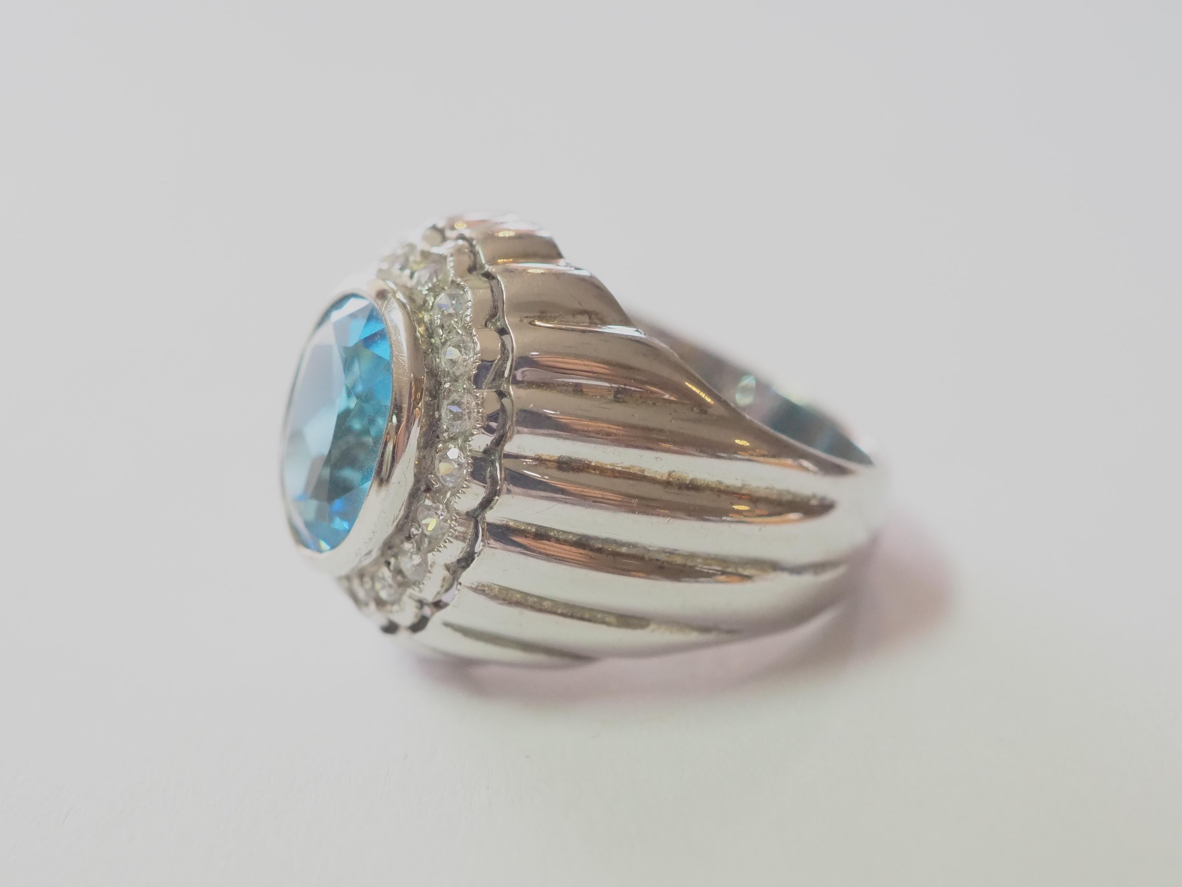This ring is a statement signet Trombino ring in solid sterling silver. The ring is decorated by natural oval blue topaz bezel set in the middle. The surrounding white stones are cubic zirconia. The engraving pattern on the band is delicately carved