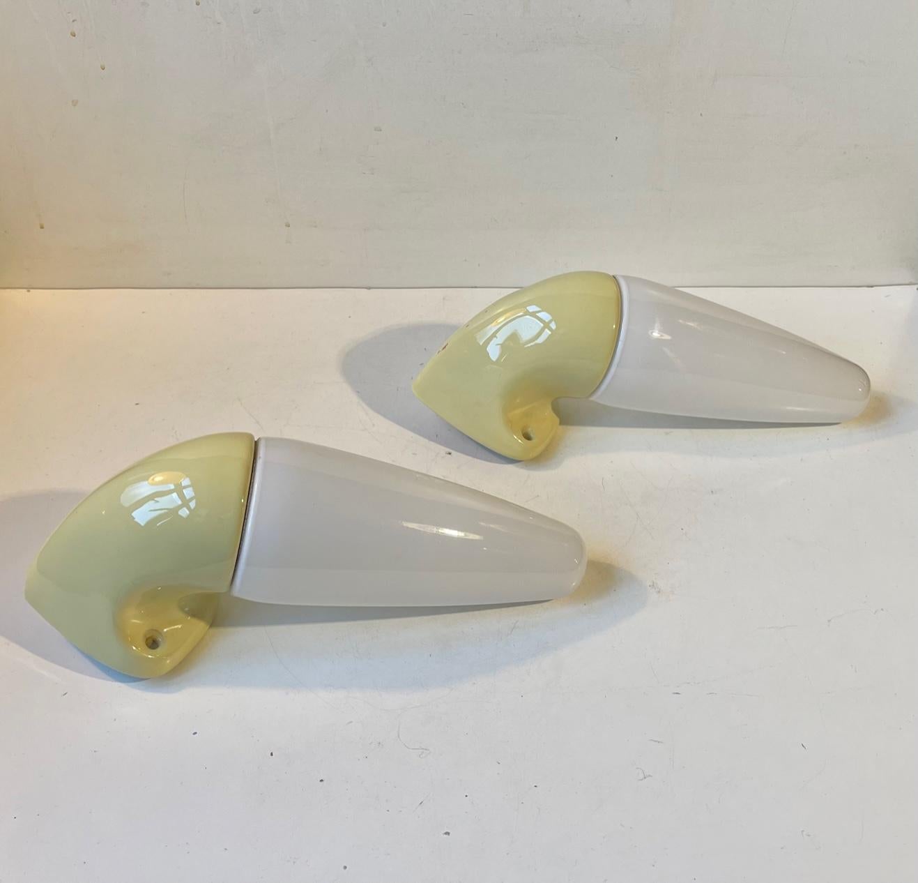 A pair of sconces suitable as bathroom or outdoor lighting. They were designed by Prince Sigvard Bernadotte for the swedish company Ifö during the 1960s. Pastel yellow glazed ceramic mounts with white opaline glass shading. 2 way mount - either