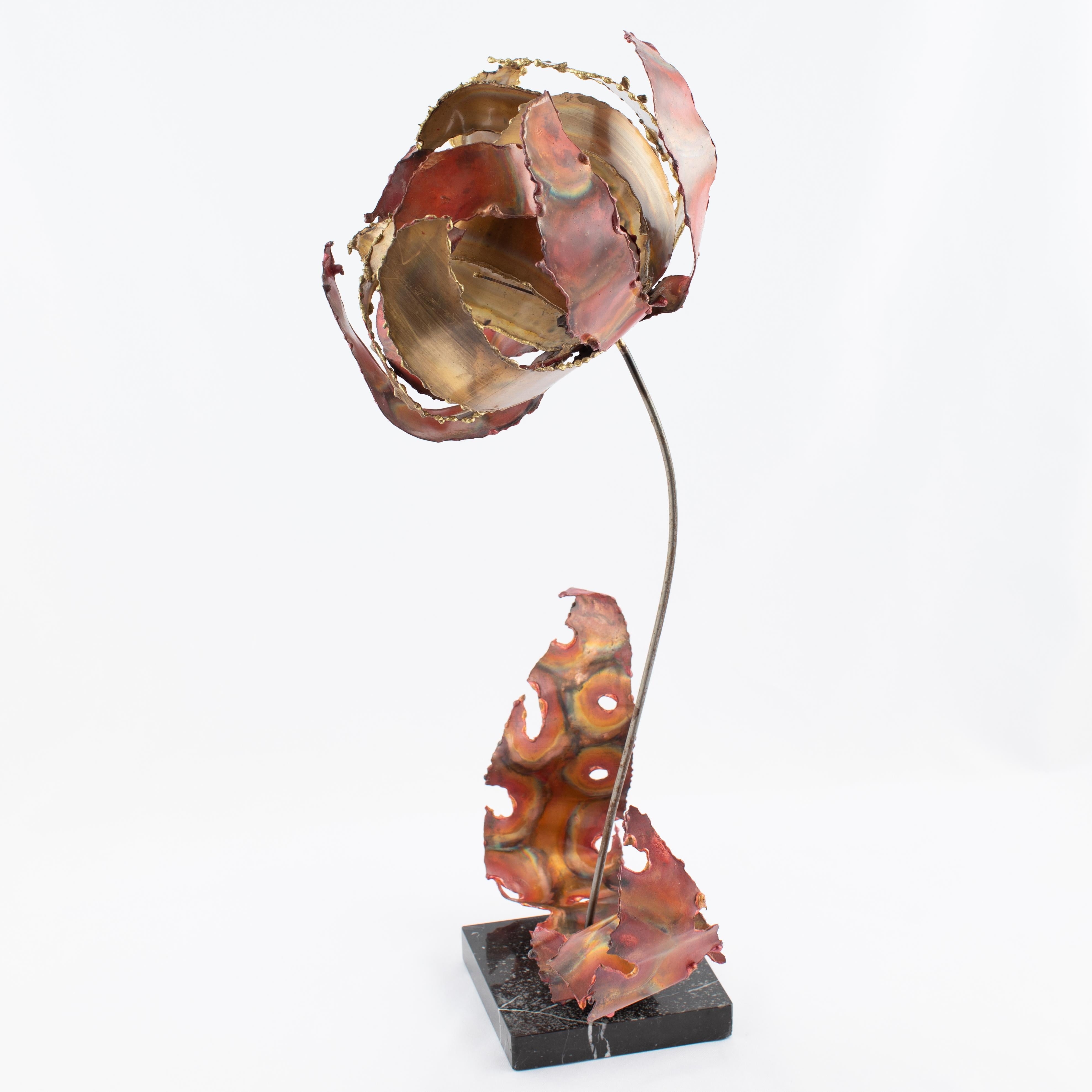 Whimsical metal flower sculpture by New York artist Silas Seandel features petals and leaves of braised copper and brass welded to a steel stem supported by a black marble base. The colors and variegated patterns result from heat and acid