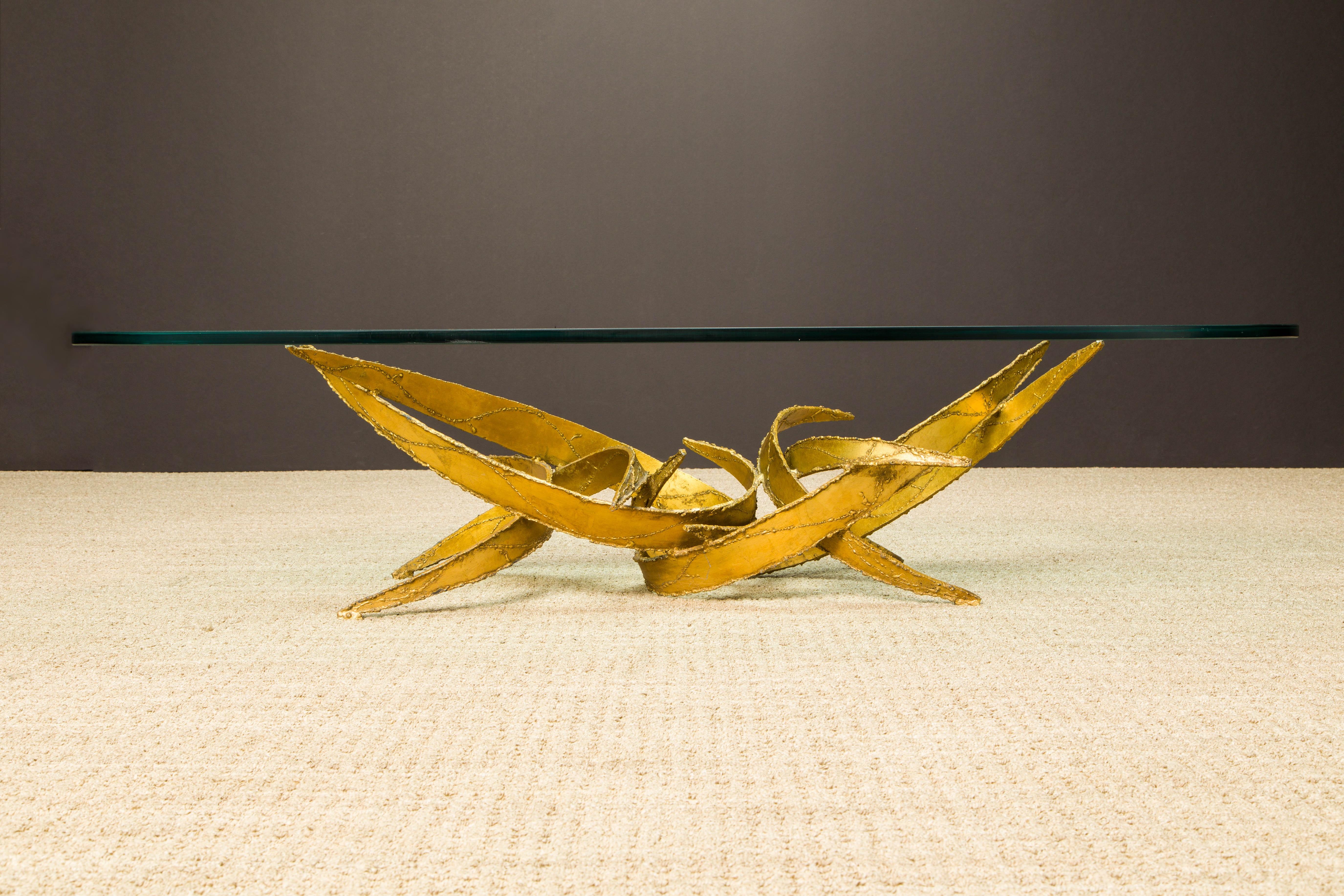 This sculptural Silas Seandel Brutalist coffee table is constructed of sections of curved gold tone torch cut metal which have been formed into an organically abstract structure and functional art piece. This structure balances a piece of glass to