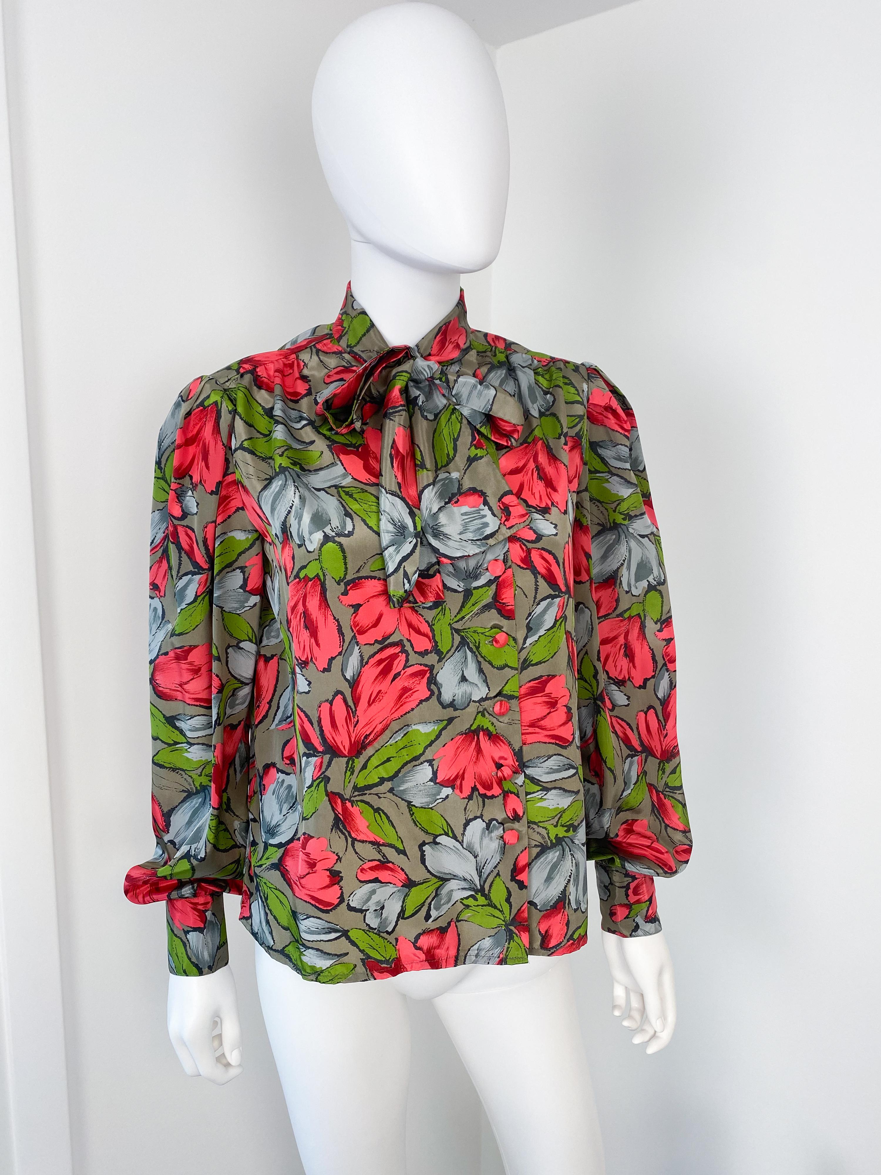 This exquisite vintage 1980s pussy bow blouse is crafted from silky viscose-polyester, radiating both elegance and charm. The taupe-colored fabric is beautifully adorned with bold floral patterns in striking shades of red, green, and gray,