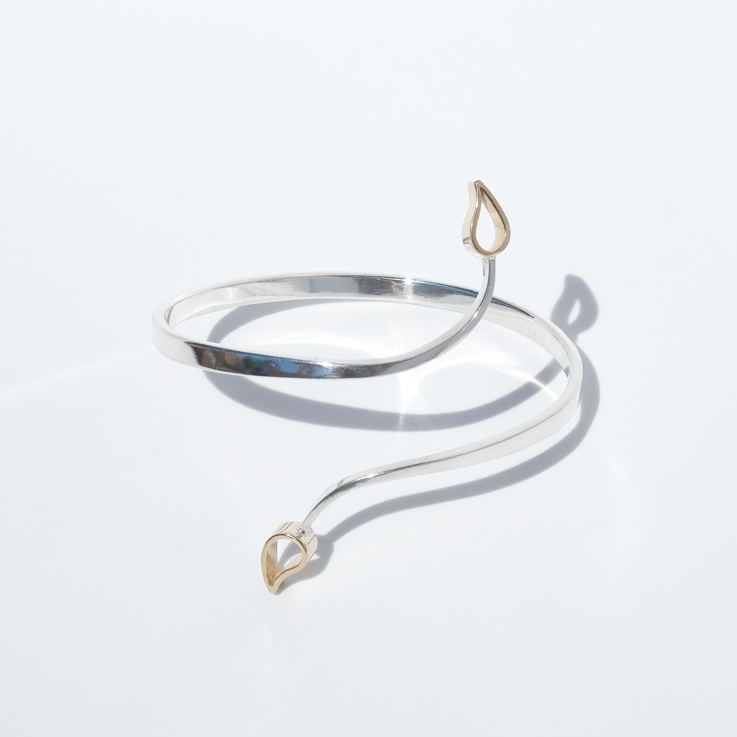 Questo bracciale in argento ha un design aperto e fluente che si avvolge con eleganza intorno al polso. La sottile fascia d'argento si curva in modo organico e termina con due delicati dettagli a forma di goccia in oro 18 carati che aggiungono