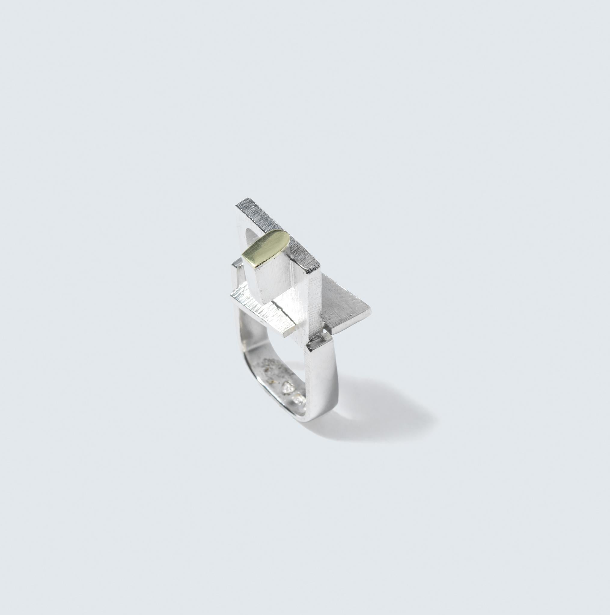 This is a unique ring that blends silver and 18 karat gold in an industrial design. The band of the ring is square in shape, giving it a modern and structured look. At the center, it features an assembly of various geometric shapes, adding to the