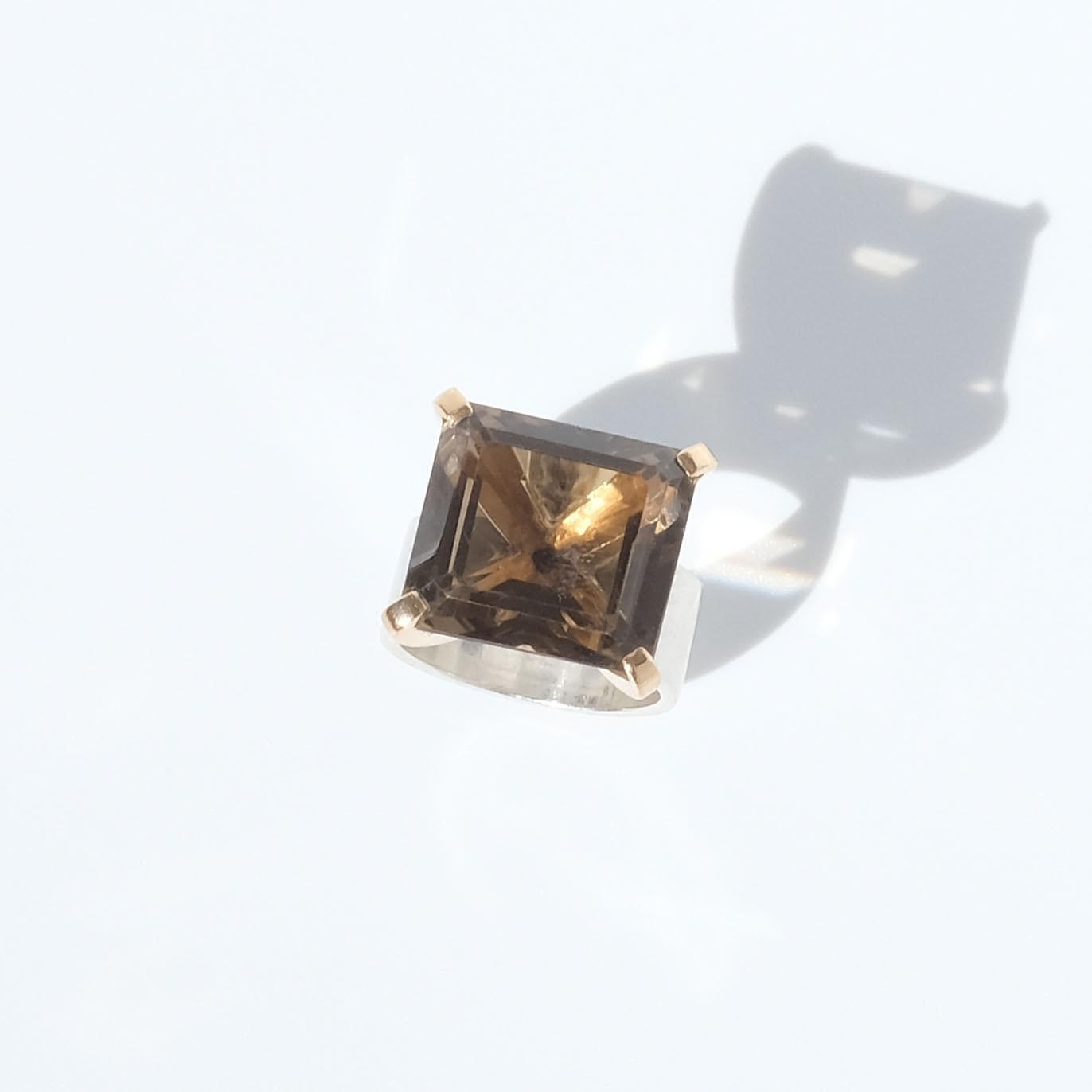 This sterling silver ring is adorned with a large, square, faceted smoky quartz stone. The head of the stone is distinct and its gilded prongs prominent. The shank of the ring. made of 18k gold, is beautifully wide and creates a strength in the