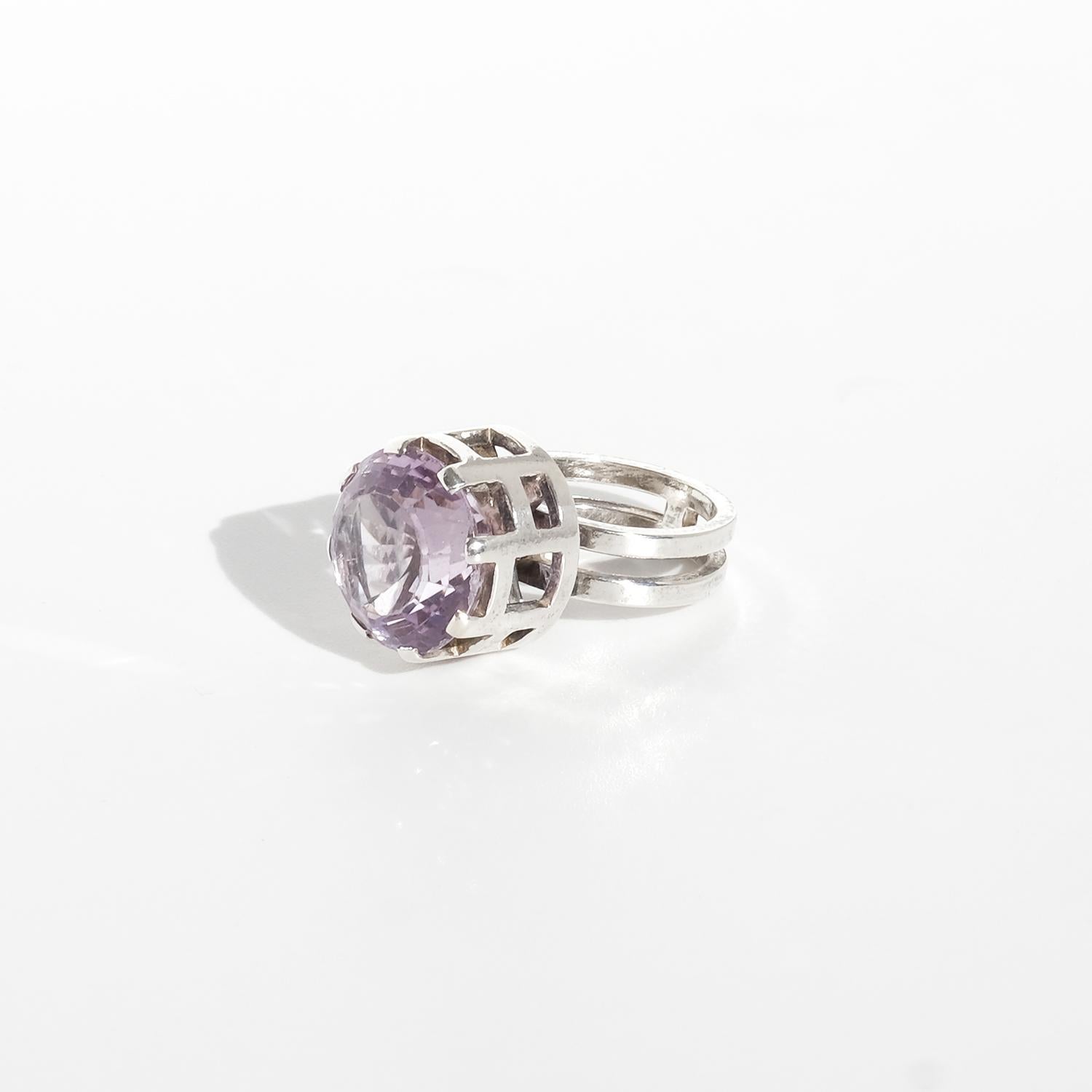 Questo anello d'argento vintage presenta un'ametista color lavanda chiaro incastonata in un castone traforato. La pietra è rotonda con una corona alta e sfaccettata che riflette la luce grazie alla sua morbida tonalità viola. La montatura è