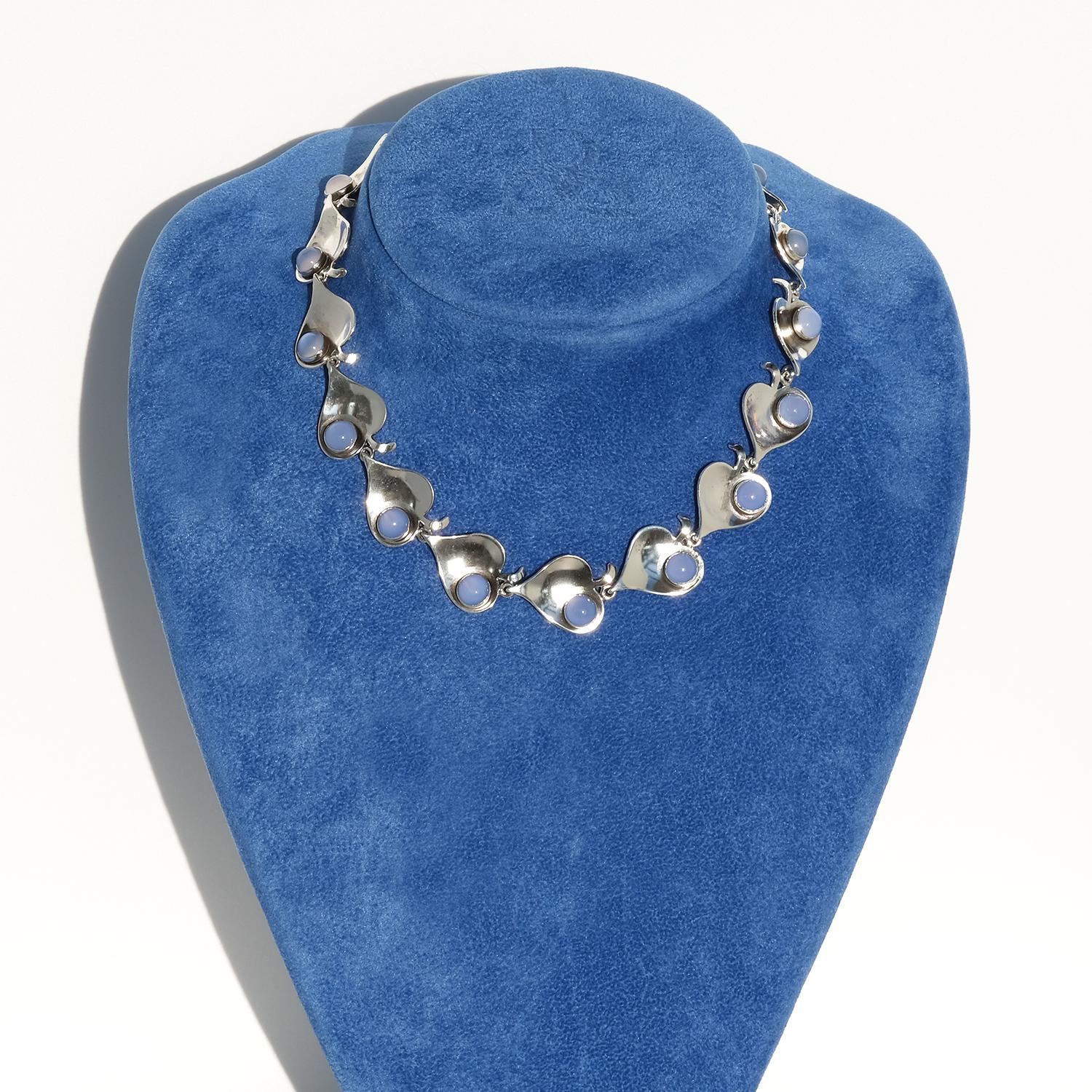 This silver choker necklace is constructed with a series of linked, heart-shaped silver segments, each adorned with a dove-blue chalcedony cabochon. The chalcedony stones display a soft, muted blue tone that contrasts beautifully with the polished