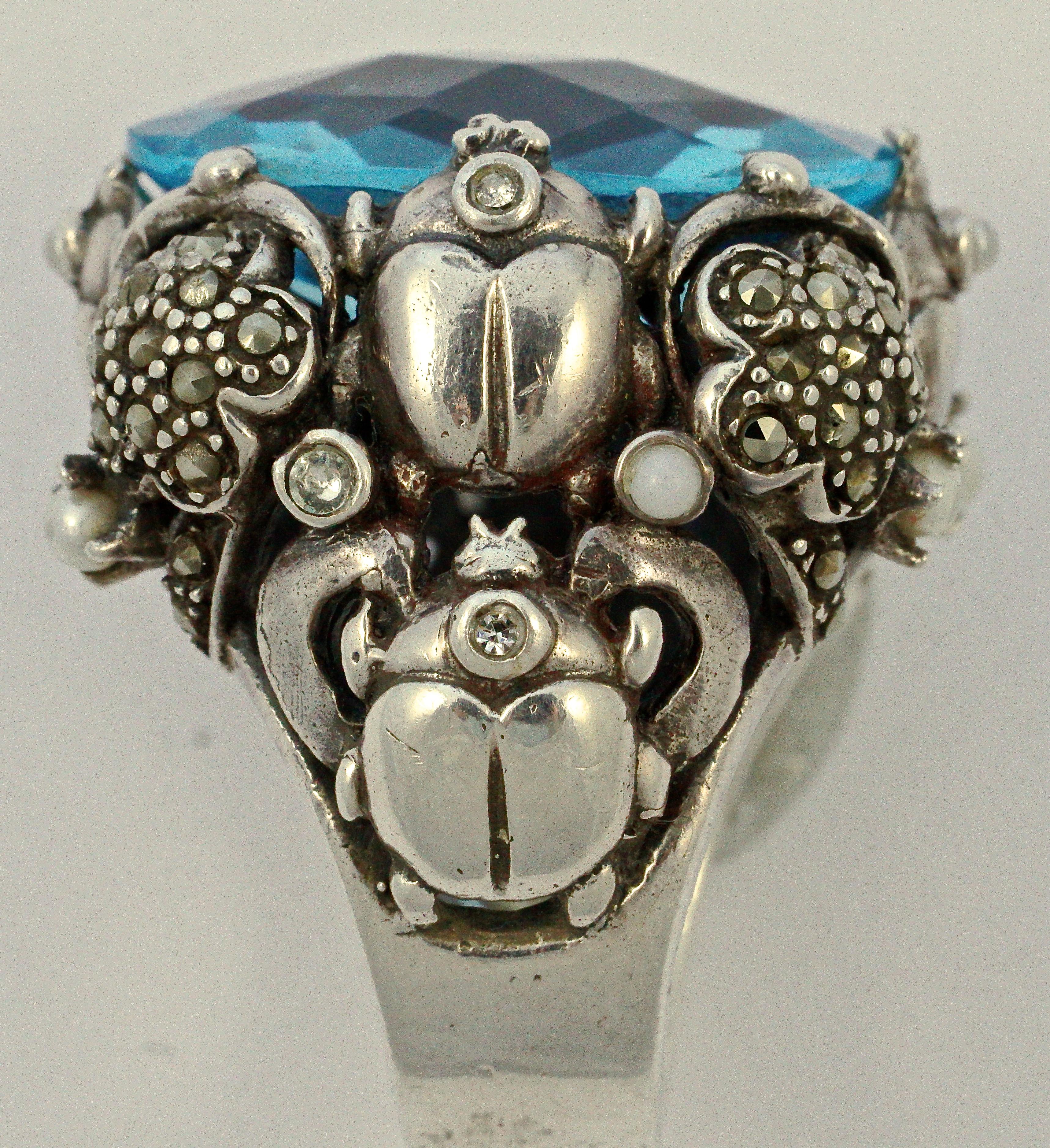 Fabulous silver ring featuring an oval faceted blue glass stone, circa 1970s. The setting has an intricate design with marcasite set flowers, and ladybirds decorated with faux pearls and clear rhinestones. Ring size UK M 1/2, US 6 1/4 (inside