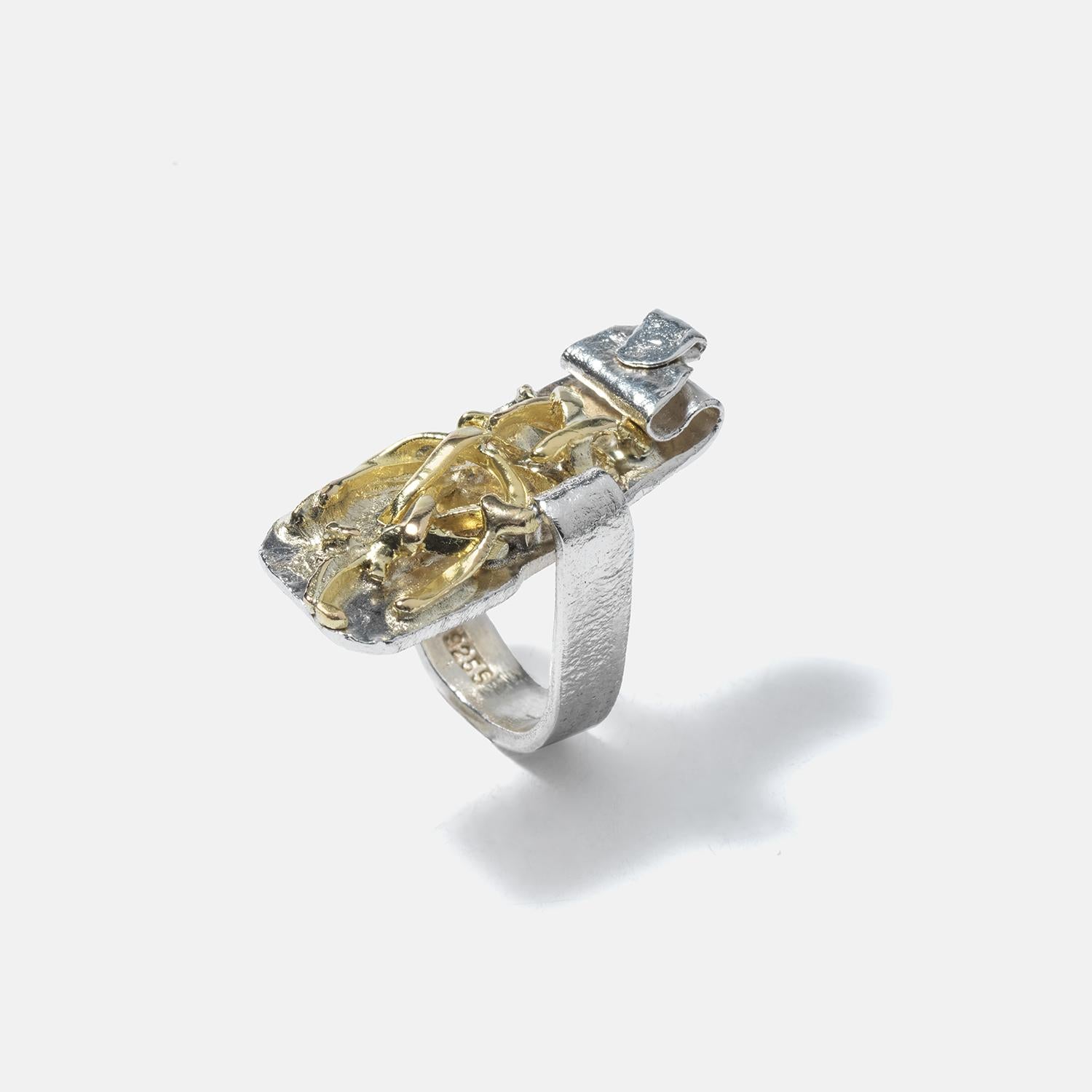 This ring is a unique piece of jewelry that combines both silver and gilded silver elements. It has a broad, unevenly textured band that narrows slightly at the back, giving it a rustic and handcrafted appearance. The top of the ring features an