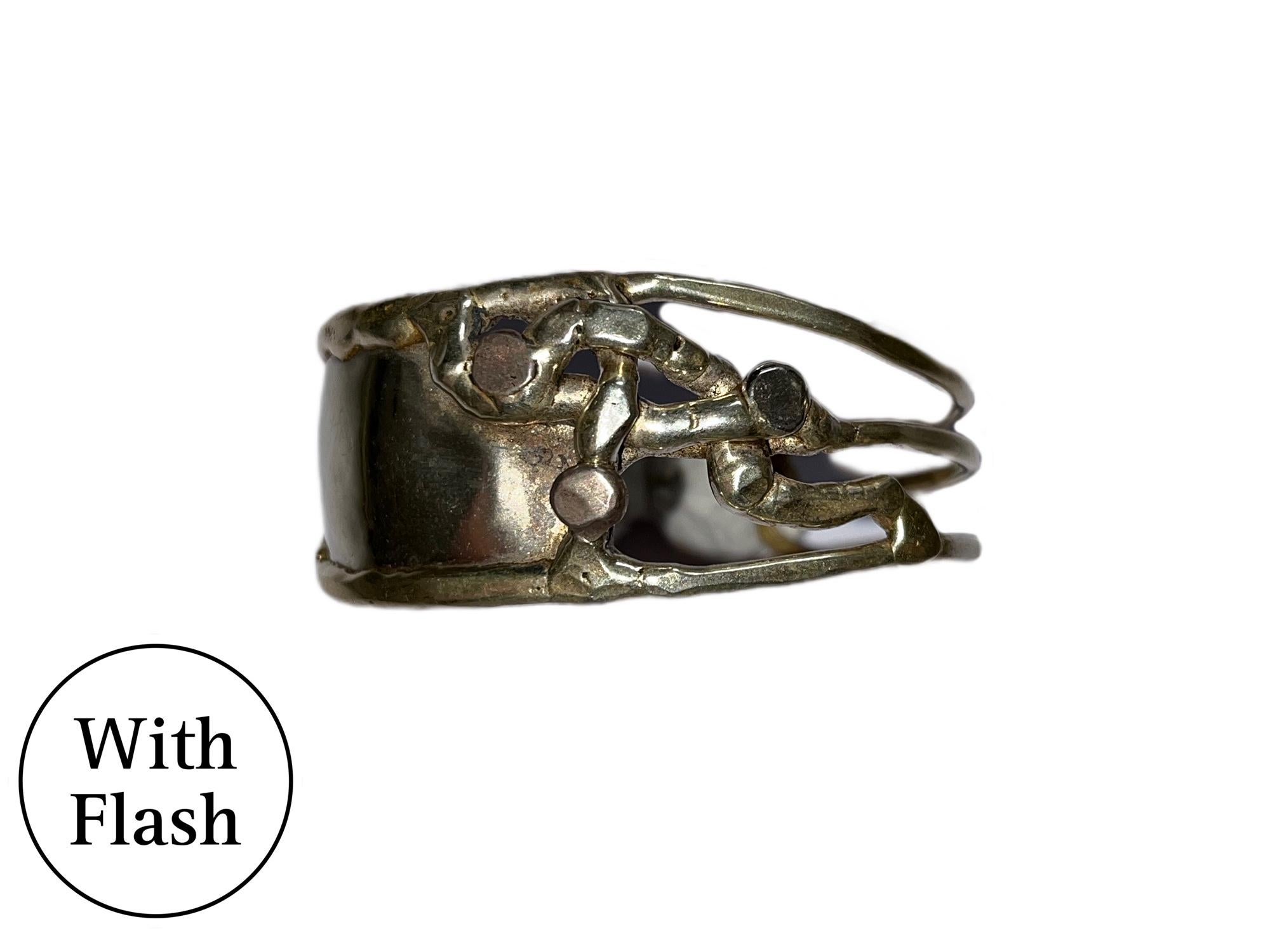 Si tratta di un braccialetto bangle in argento e oro dal design moderno e scultoreo. Il bracciale è caratterizzato da un'ampia fascia di metallo lucido che avvolge il polso solo per metà. Il dettaglio di spicco di questo pezzo è l'intricata