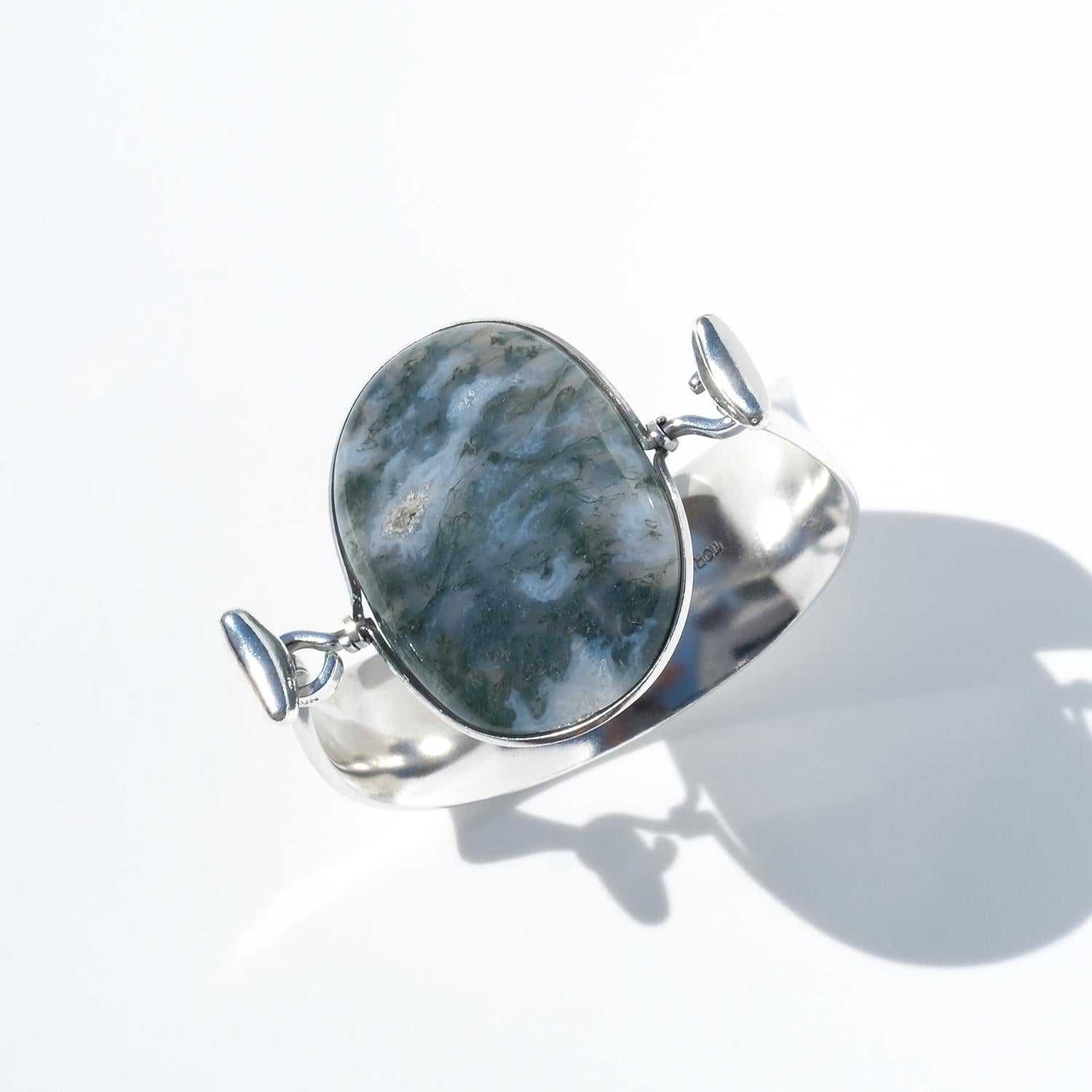 This bracelet is crafted in silver and features a wide, slightly concave band that tapers gently toward the top, creating an elegant upward sweep. The ends of the band curve inward, each finished with a softly rounded oval form. At the center sits a