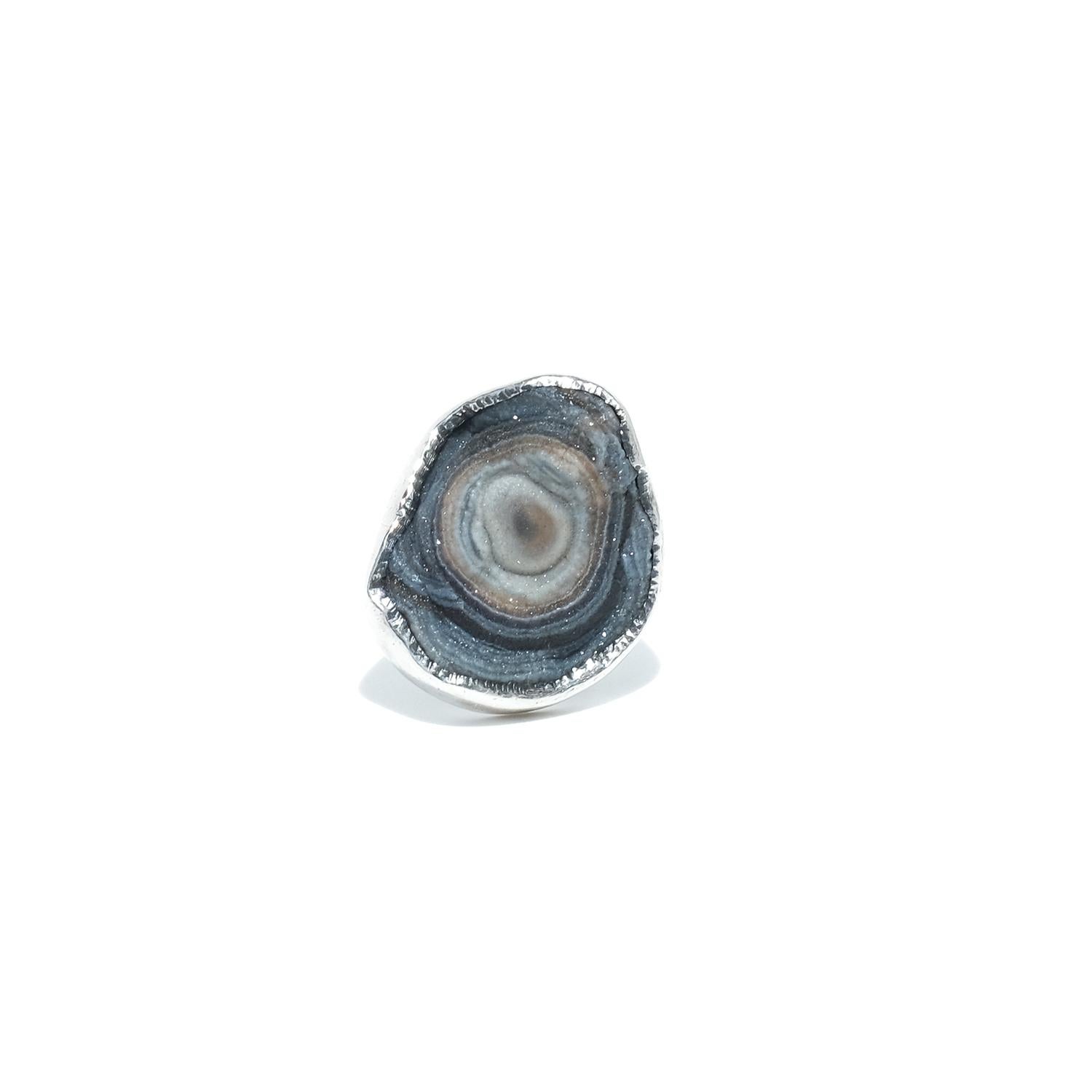 This silver ring features a large, three-dimensional natural stone with concentric layers of gray, black, and beige tones. The wide bezel setting is irregular in shape, carefully crafted, and textured along the edges. The stone is prominently raised