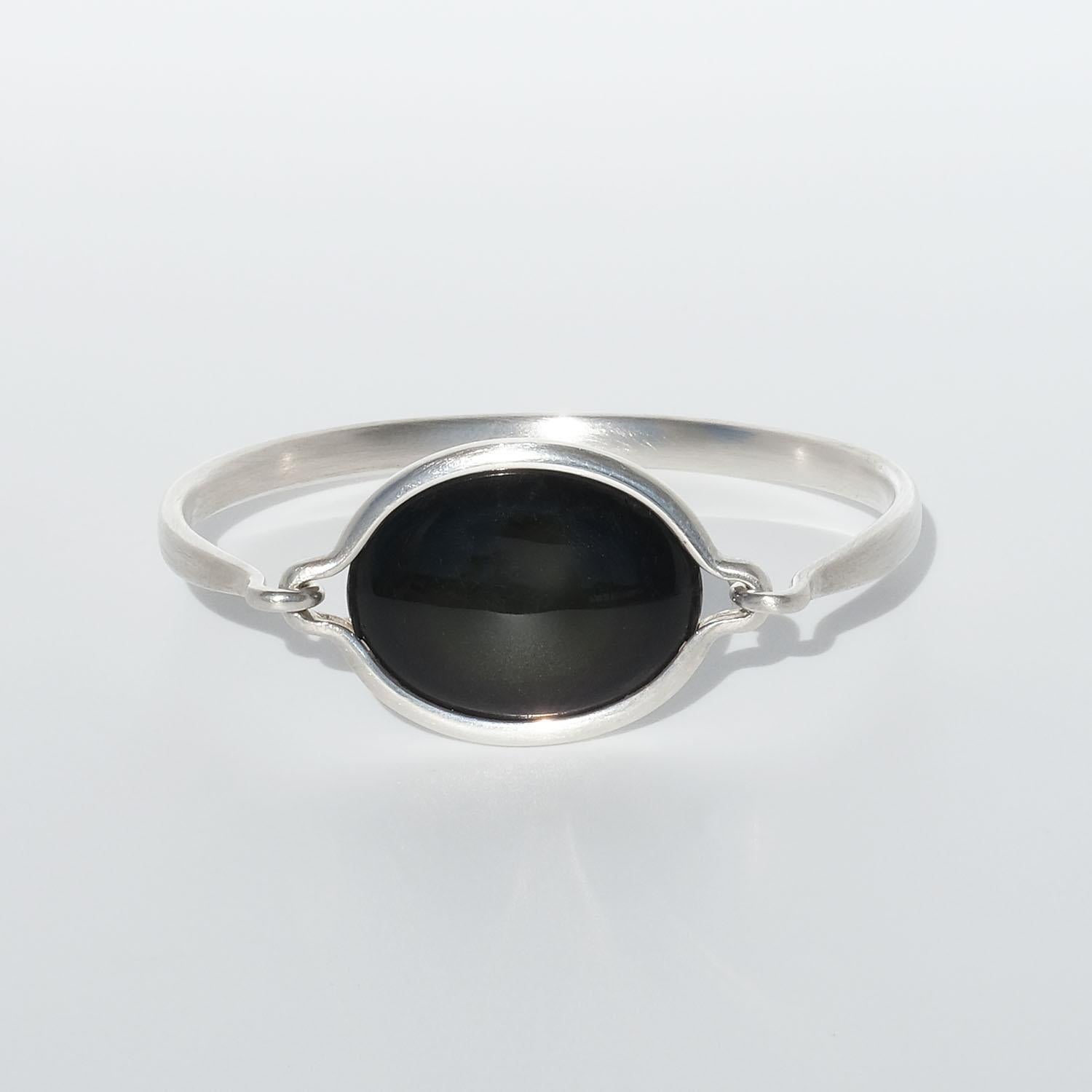 Dieser Armreif aus Sterlingsilber ist mit einem ovalen, glänzenden Regenbogenobsidian verziert. Der Obsidianstein ist mit zwei Haken am Armband befestigt und kann abgenommen werden. Im Regenbogenobsidian haben sich mikroskopisch kleine Blasen