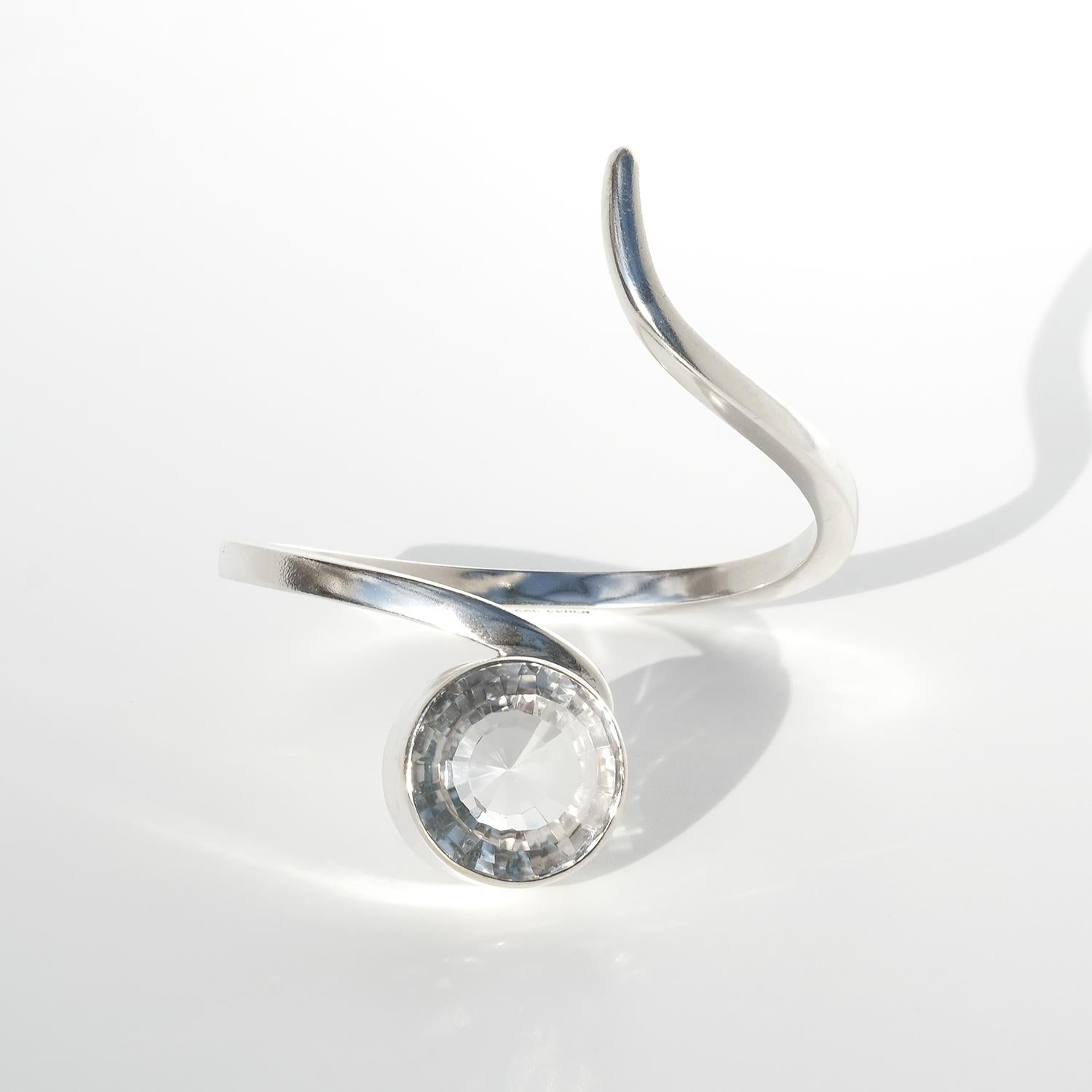 Dieses silberne Manschettenarmband zeichnet sich durch ein asymmetrisches Design mit einer anmutigen, gewundenen Form aus, die sich um den Arm wickelt. In seiner Mitte befindet sich ein großer, facettierter Bergkristall, der in einer hohen,