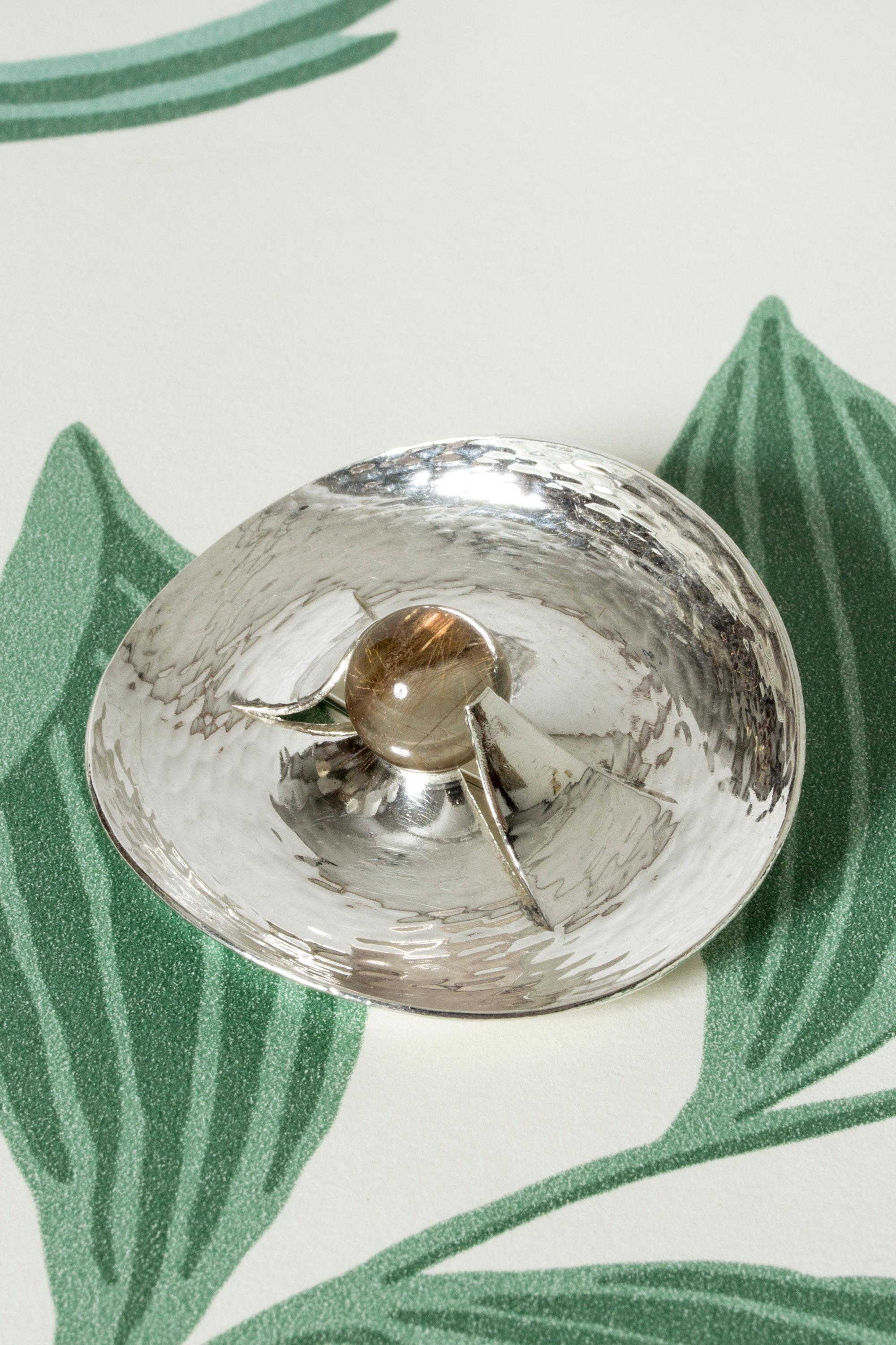 Amazing silver brooch by Elis Kauppi, in an oval form with a hammered surface that creates a rippling effect. A round rutilated quartz stone in the center seems to be emerging out of the silver.