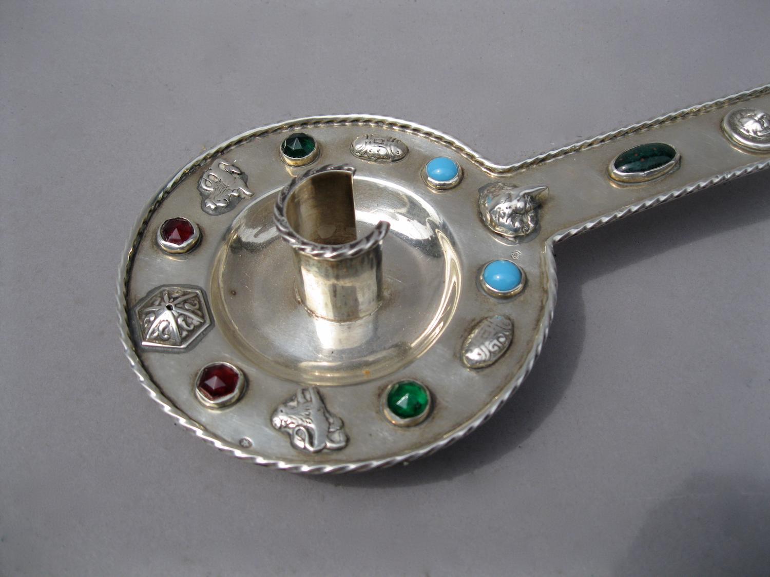 Circular silver candleholder, standing on two small legs and extended by a gripper.
The whole ensemble is decorated with lion heads, lying heraldic lions, a shell, and geometrical medallions, alternating with colorful stones in turquoise,