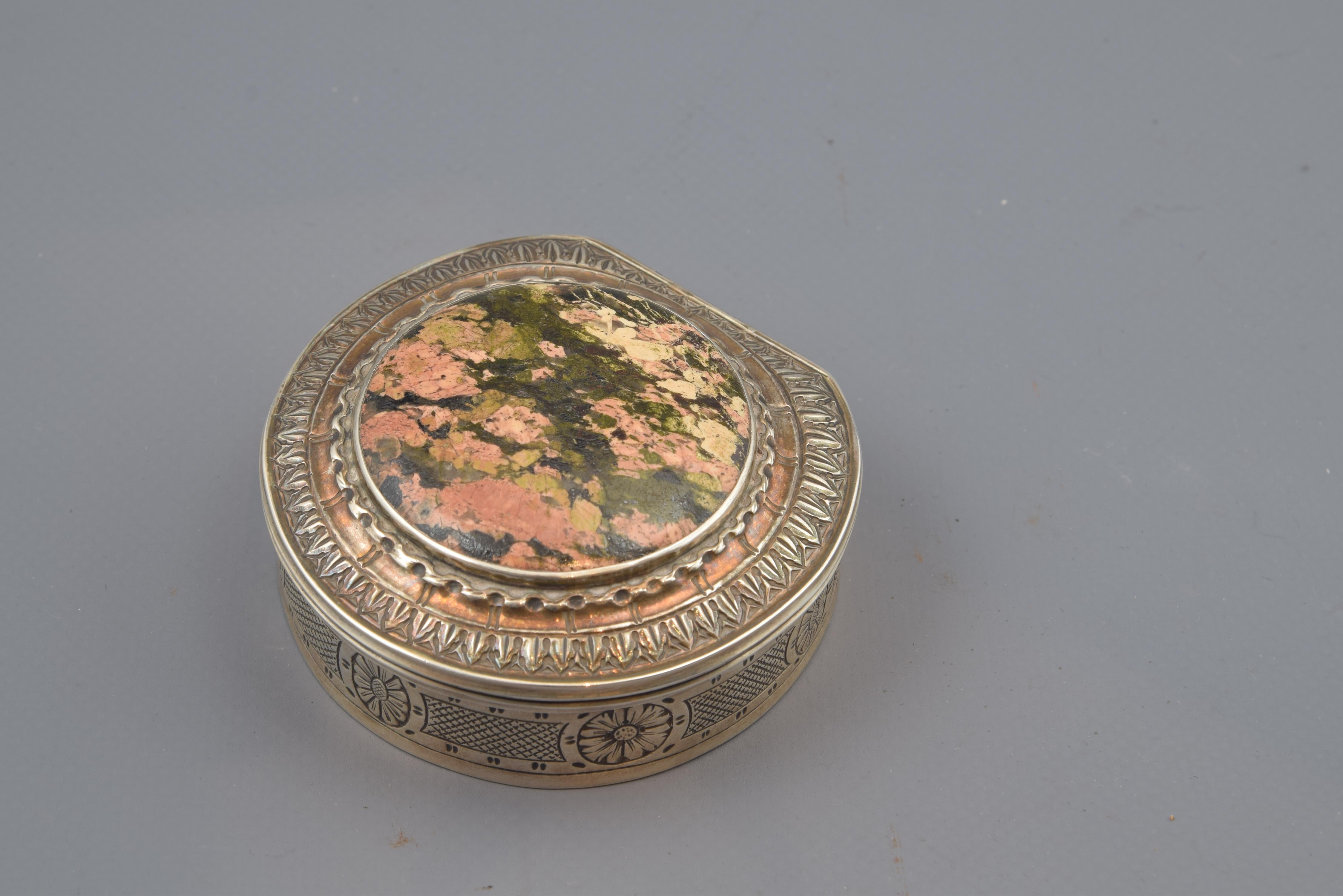 With hallmarks.
Silver box in its almost circular shape with the border decorated with flowers in a band of textile-like lines. The cover has a series of bands of plant elements and others that enhance a polished stone, located in the centre of it.