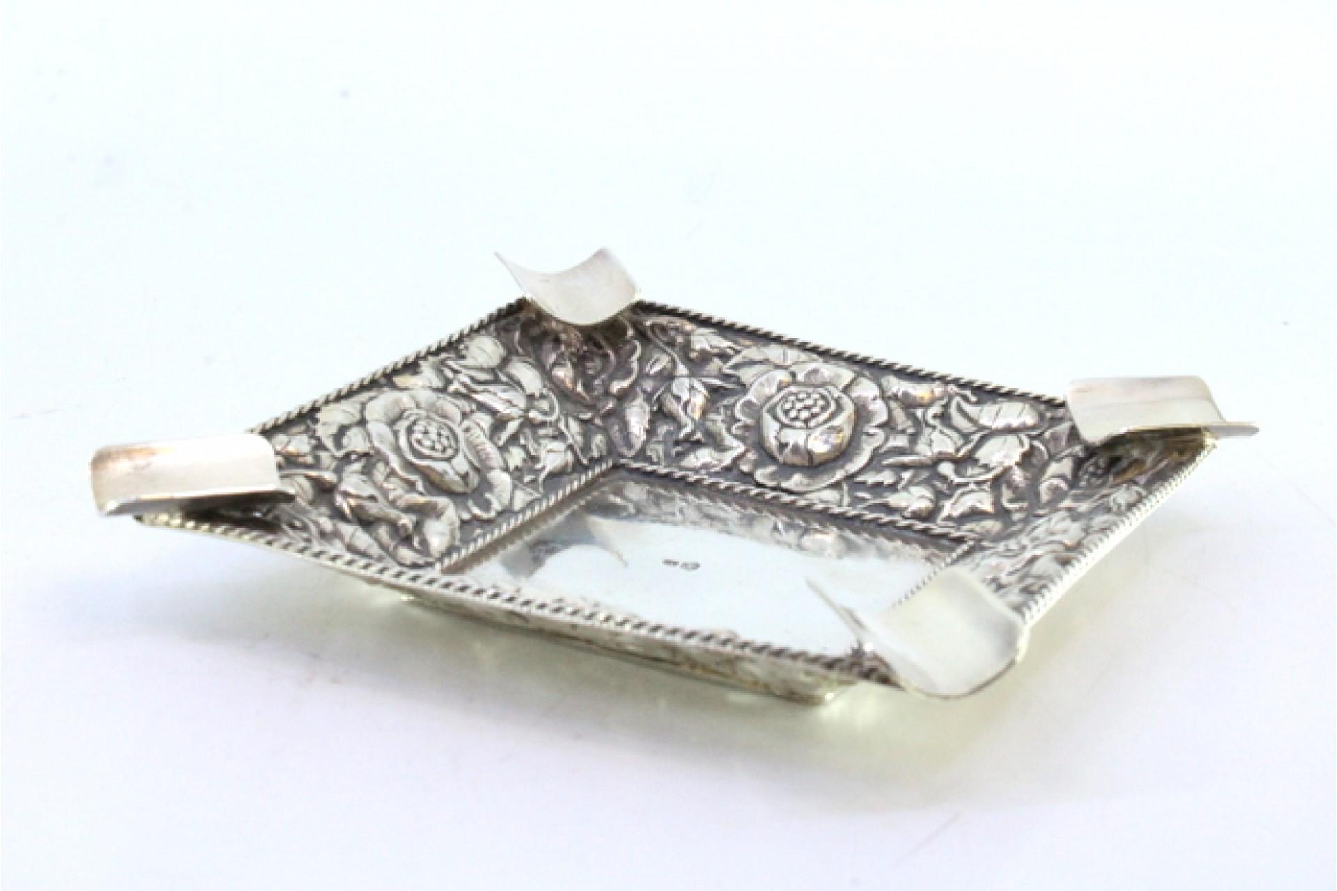 Descripción: Cenicero de plata con motivos florales en relieve, friso acordonado y cuatro pitilleras. Metal: Silver. Origen: Portugués. Marca: águila de alas abiertas. Peso: 105,57 gramos. Dimensiones: 11cm x 11cm. Contenido: 835/1000. Marca de