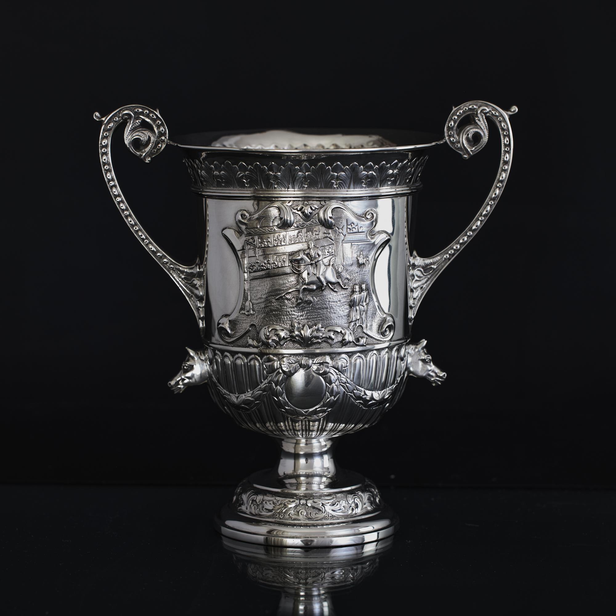 Unusual antique two-handled silver trophy cup with cast and hand chased detail including a cavalry horse riding scene depicting a competition of horsemanship inside an arena. There is further decoration of acanthus leaves, fluting, wreaths and