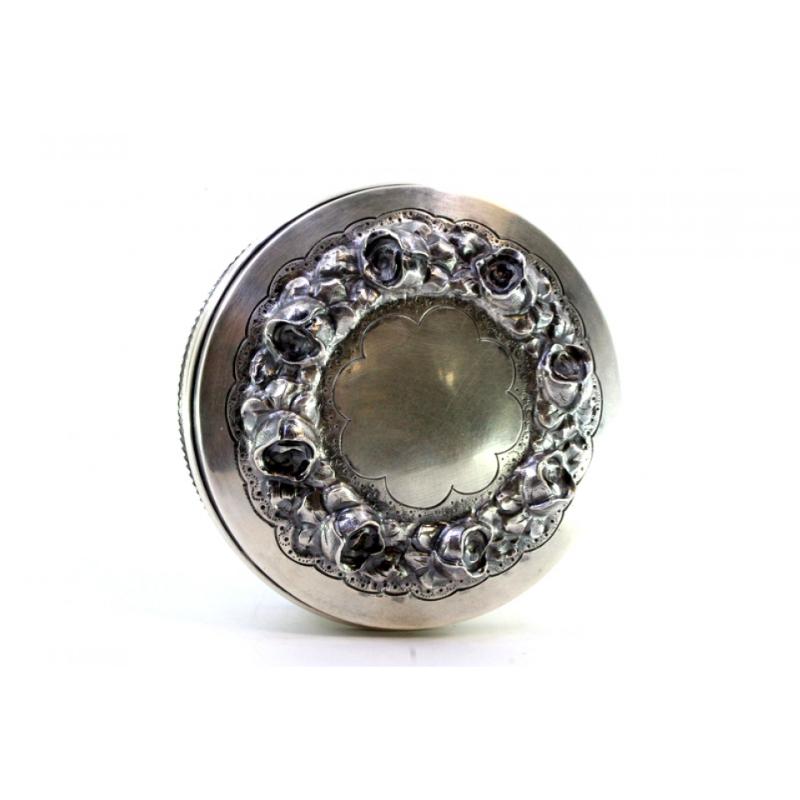 Description: Silver box with embossed floral motifs and a surrounding frieze on the circular base. Metal: Silver. Origin: Portuguese. Mark: standing eagle. Weight: 118.75 grams. Dimensions: 10cmx4cm. Content: 833/1000. Assay Mark: Yes, between 1938