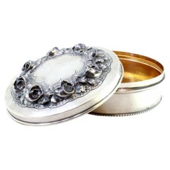Silver box w/ raised floral motifs & a surrounding frieze on the circular base