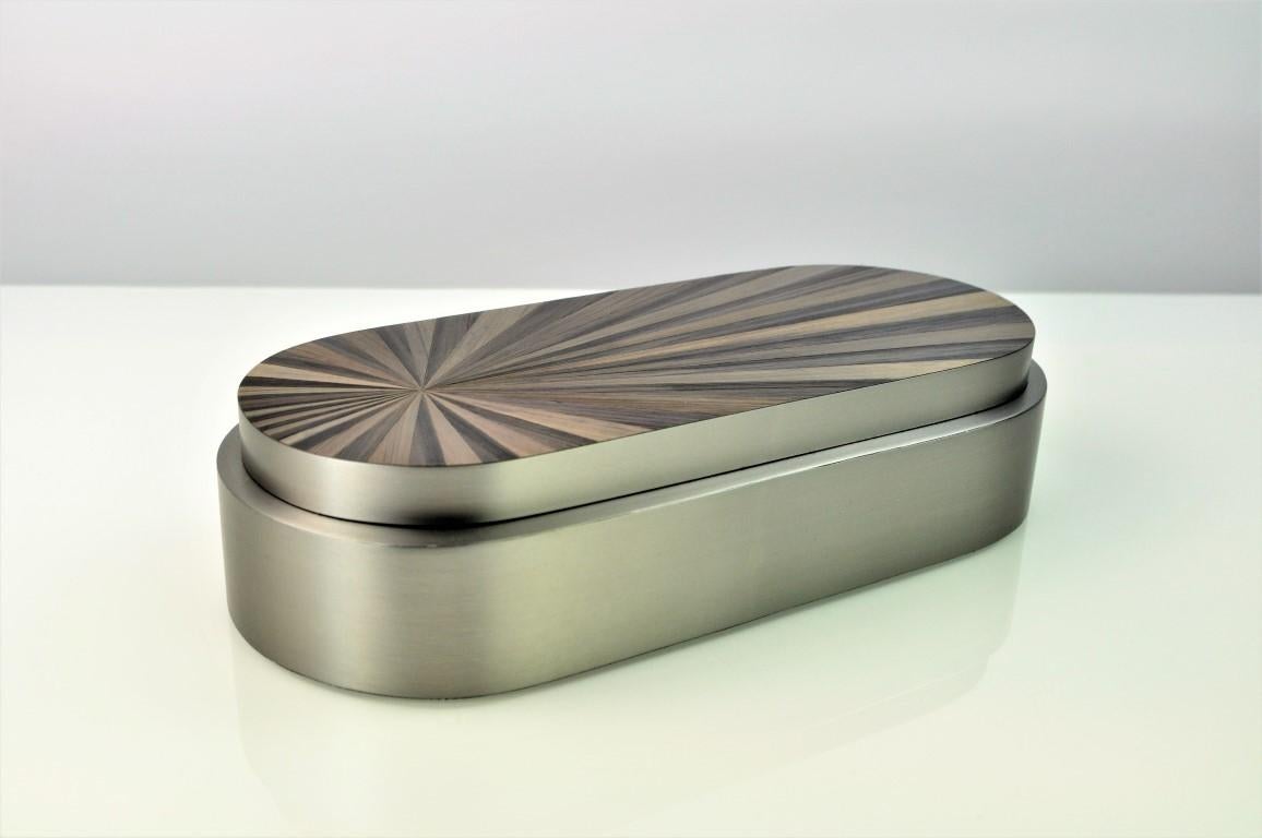 Esta preciosa caja está hecha de acero inoxidable cepillado con una tapa recubierta de marquetería de paja. 
El diseño radial de la paja recuerda al gran periodo Art déco, que contrasta perfectamente con el acabado en acero satinado.
La base tiene