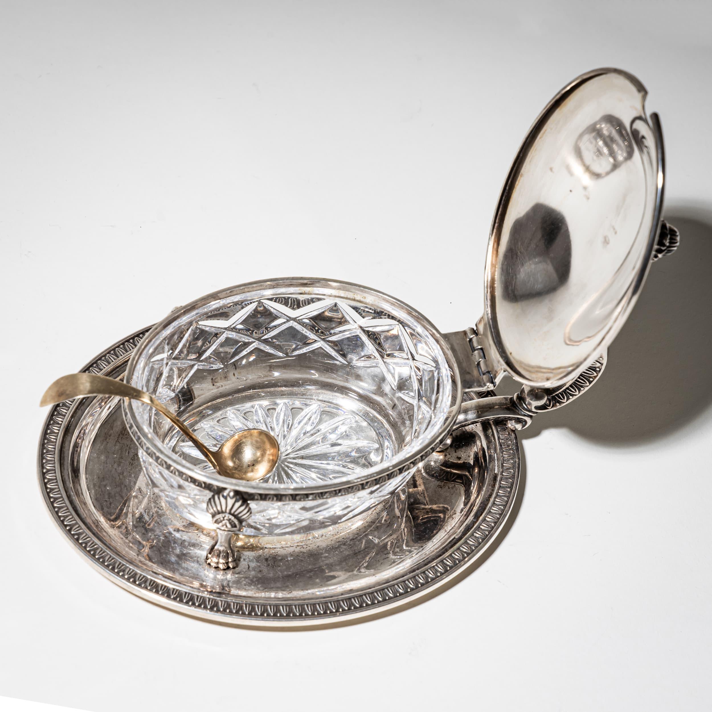Boîte à couvercle ovale avec insert en verre facetté, reposant sur quatre pieds en patte de lion et posée sur un plateau assorti. Le couvercle est orné d'un fleuron, tandis que la poignée est formée de gracieuses volutes C.C. Le couvercle et le