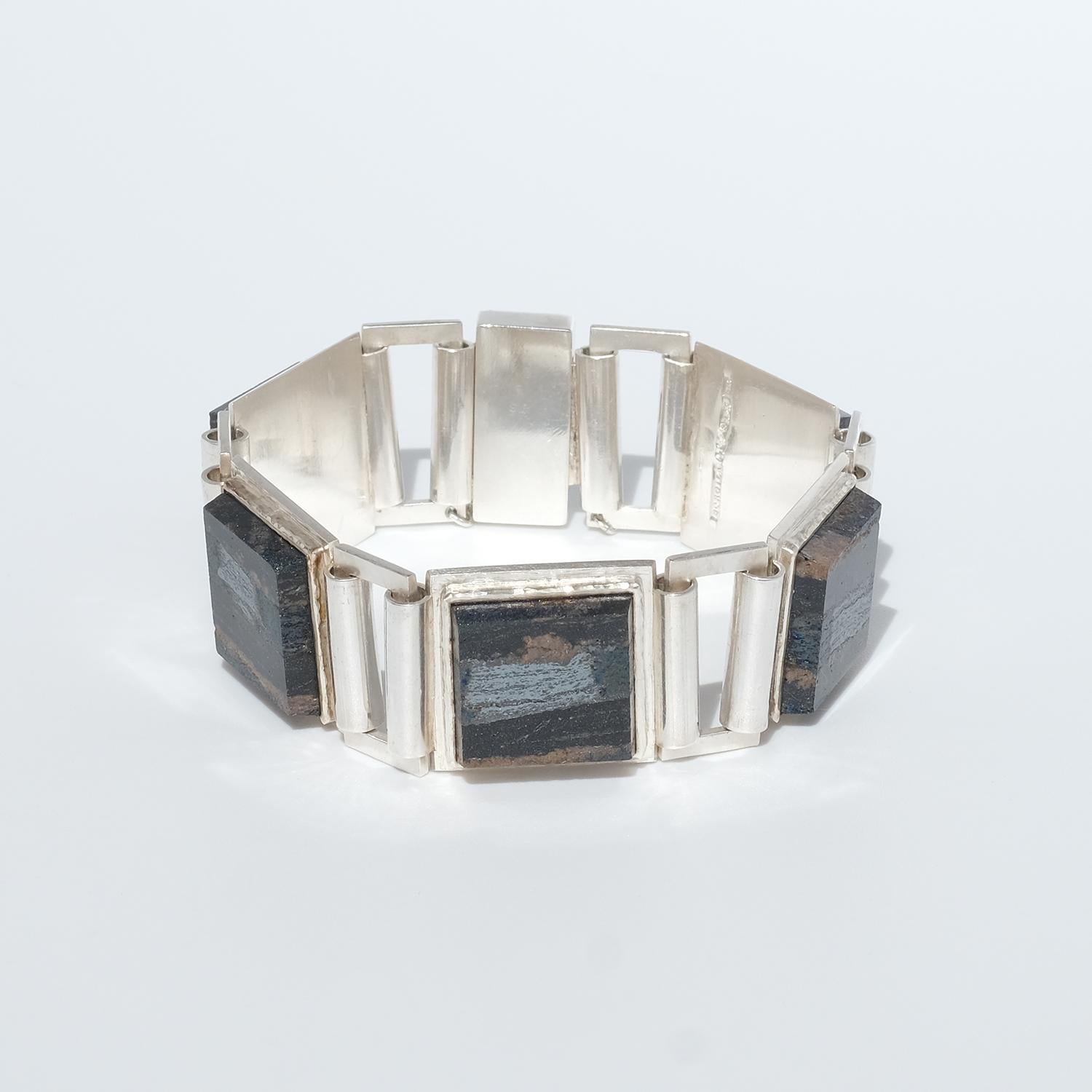 Ce bracelet est composé d'argent sterling et de cinq pierres ardoises carrées. Les pierres d'ardoise sont reliées entre elles par des rectangles d'argent. Le bracelet se ferme facilement grâce à un fermoir en forme de boîte qui possède une chaîne de
