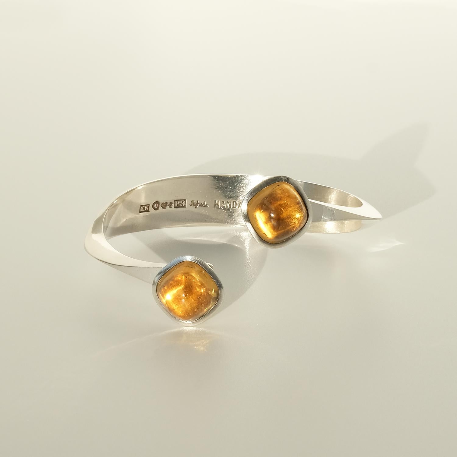 This silver cuff bracelet is adorned with two golden brown citrines that are mounted on silver square barrels. Once in place, the gemstones and their barrels look like powerful bowls of fire. The silver braclet itself is twisted on each side on its