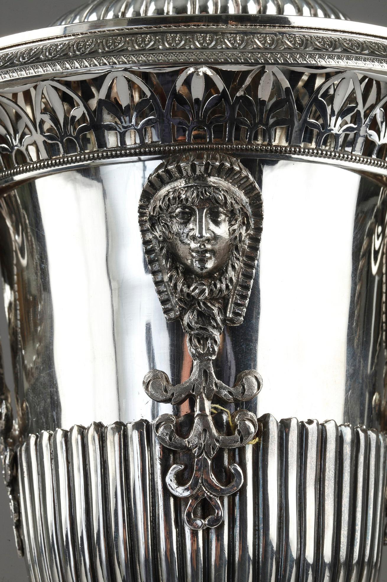 Silberne Dragee- oder Bonbonschale aus dem frühen 19. Jahrhundert, verziert mit ägyptischem Kopf und durchbrochenen Palmettenmotiven auf dem Kragen. An den beiden spiralförmigen Griffen sind zwei gedrehte Ringe aufgehängt. Der Sockel ist mit