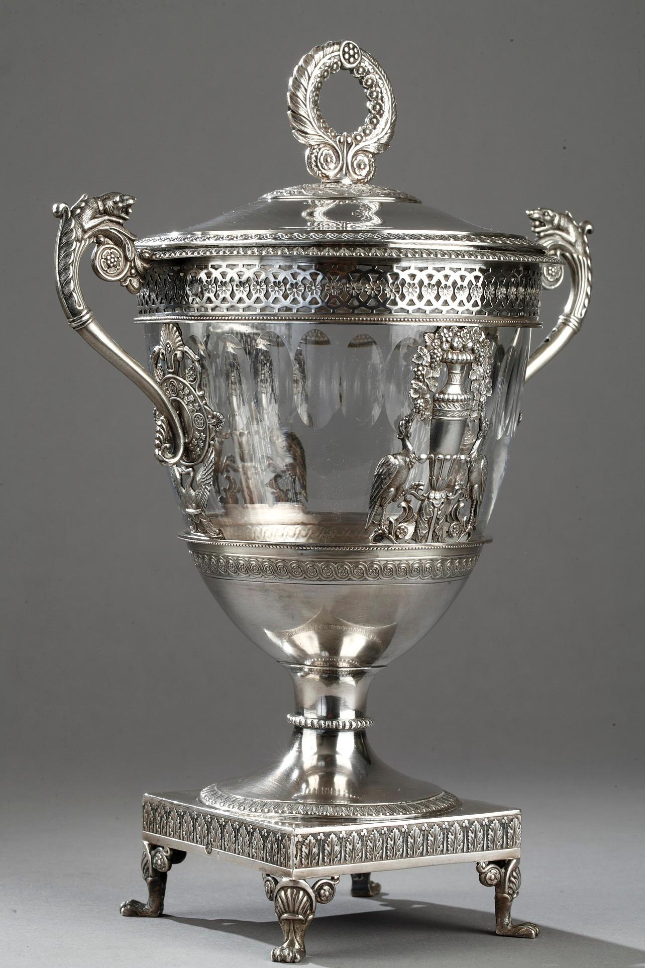 Lightly tapered candy dish in silver and crystal. The rim is ornamented with an openwork geometric frieze, and the paunch features a sculpted silver balustrade vase and two birds standing facing each other on either side of the vase. The birds are