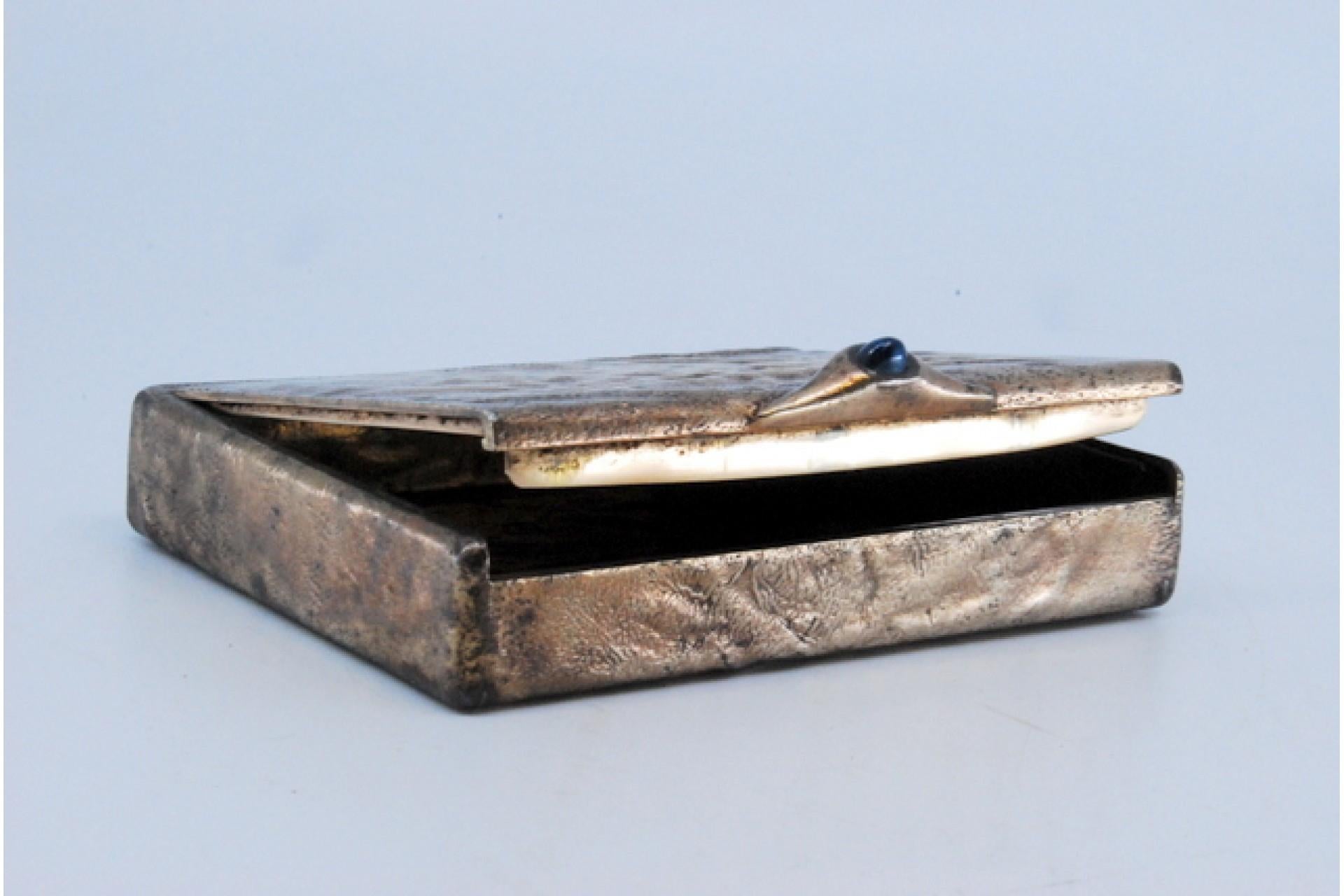 Description: Silver cigarette case with dark blue application on the lid handle and reliefs on the box. Metal: Silver. Origin: Portuguese. Brand: Cabeça de escravo. Weight: 159.91 grams. Dimensions: 9x7cm. Content: 833/1000. Assay Mark: Yes, between