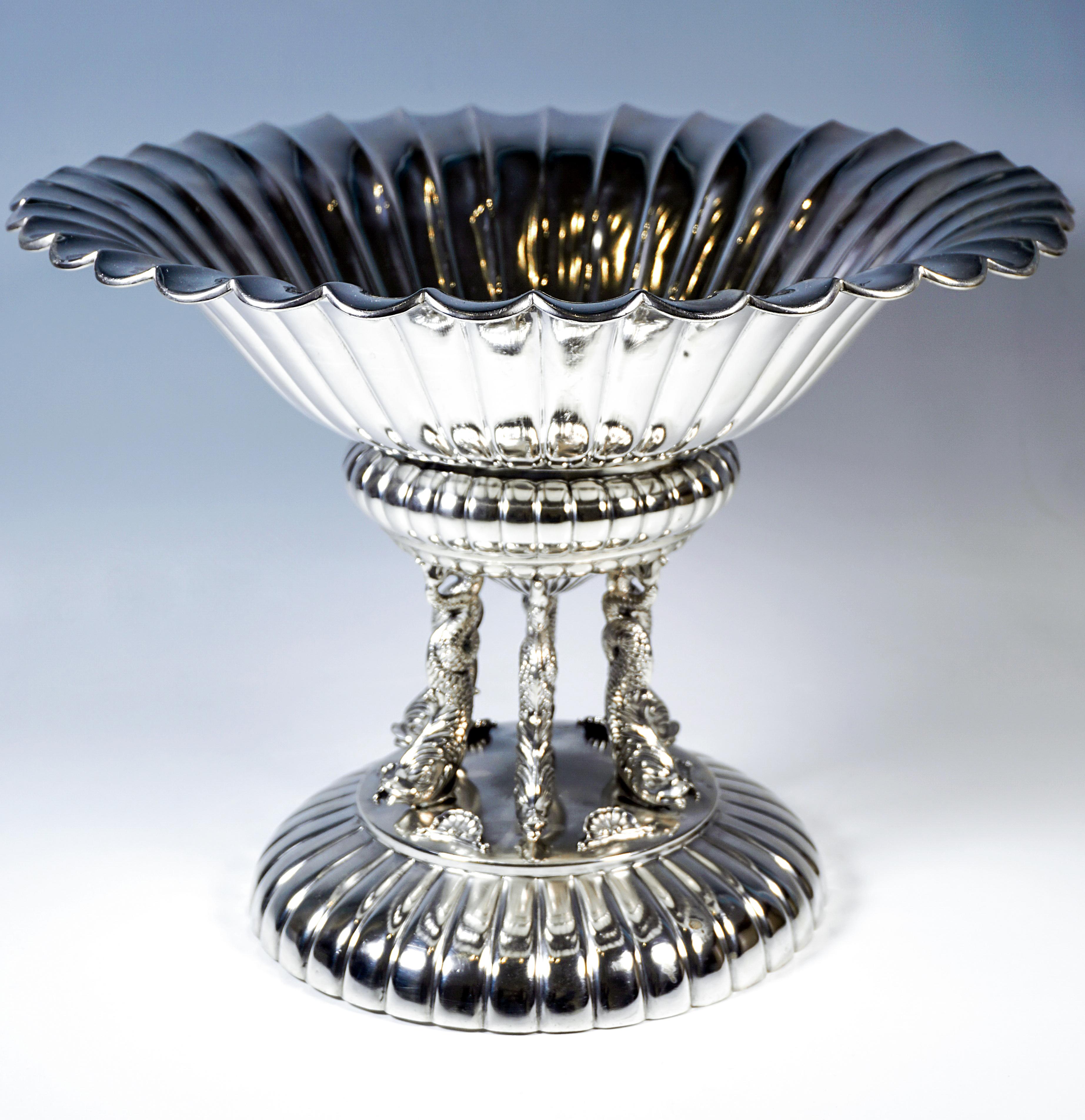 Elegante ciotola d'argento con base rotonda, che poggia su un ampio piede arrotondato a cupola con decorazione uniforme a pieghe intorno al bordo, piedistallo liscio su cui poggiano sei delfini completamente scolpiti con le pinne caudali arricciate