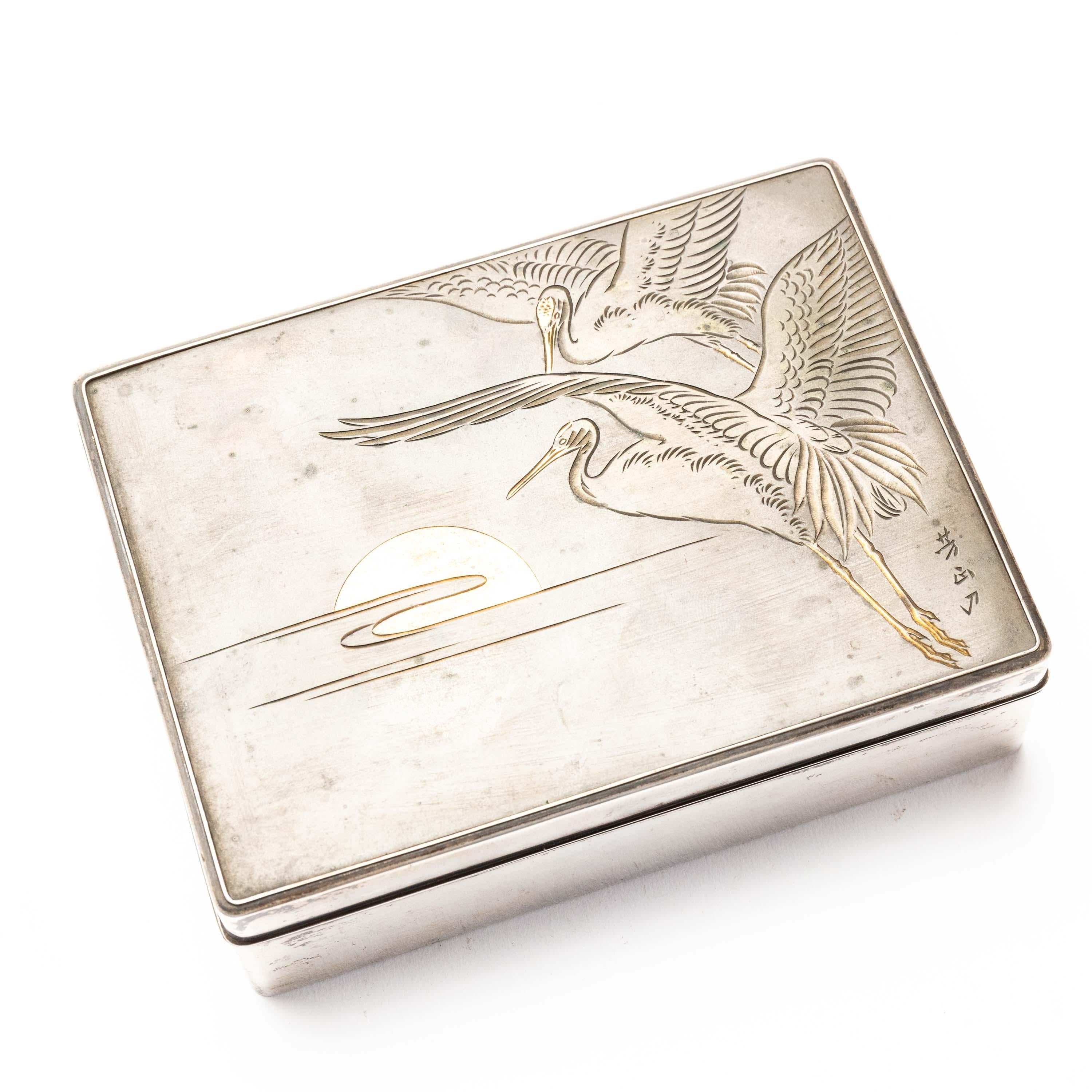 An elegant silver cigarette box from Japan, with a wooden lacquered interior. It has a delicately engraved bronze lid with a design of two cranes in flight against the rising sun. It has an artist signature on the bottom right hand corner. The