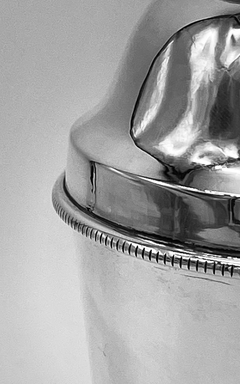 An English sterling silver cocktail shaker of plain form with a narrow Art Deco line border to the base of the lid. 
It was made by the firm of Alexander Clark & Company Limited and is hallmarked Birmingham 1926. A true classic.