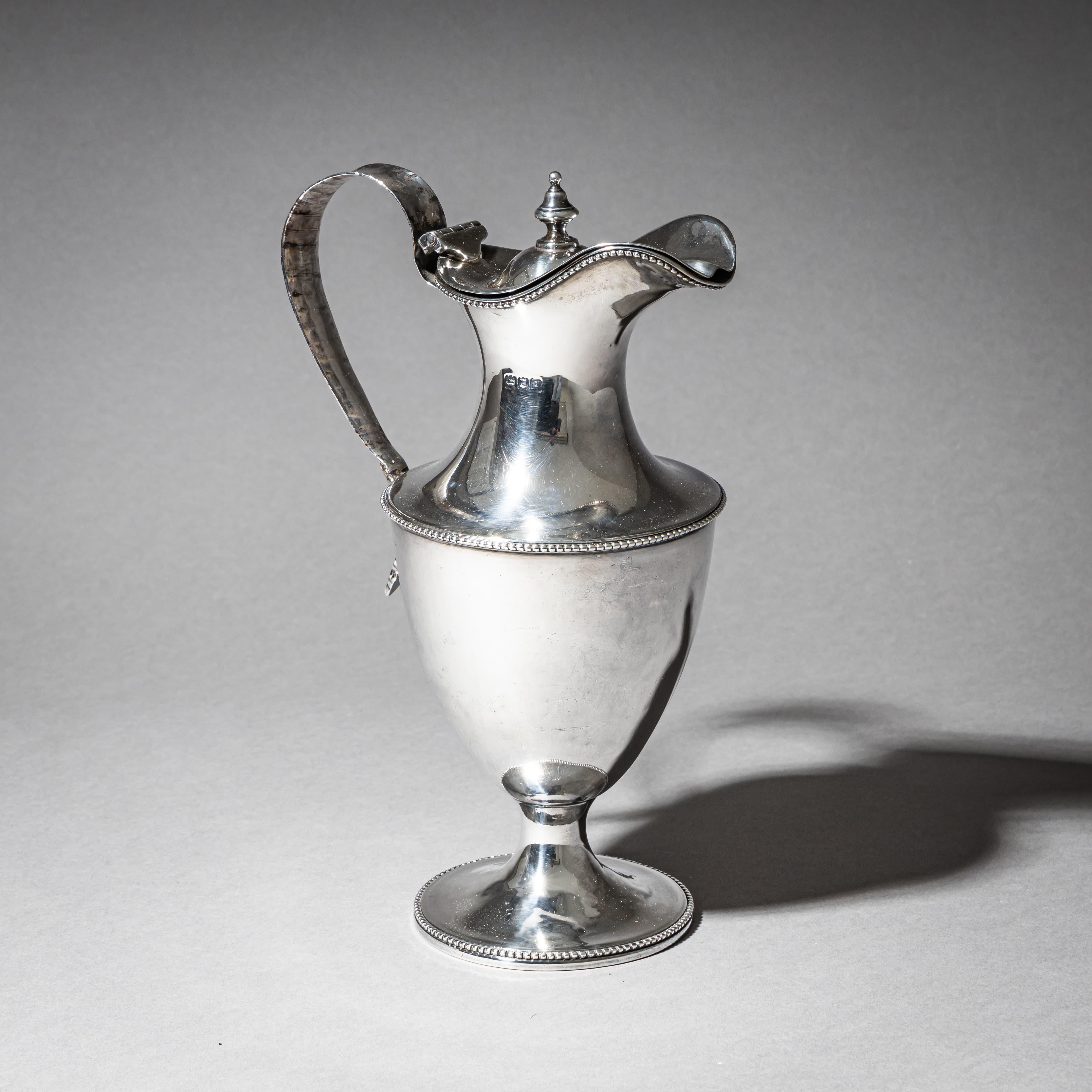 English silver coffee pot in a neoclassical urn shape, featuring a domed lid with a finely turned finial and a curved handle adorned with a leaf motif at the joint. The lid and foot are embellished with delicate beaded borders.Hallmarked on the
