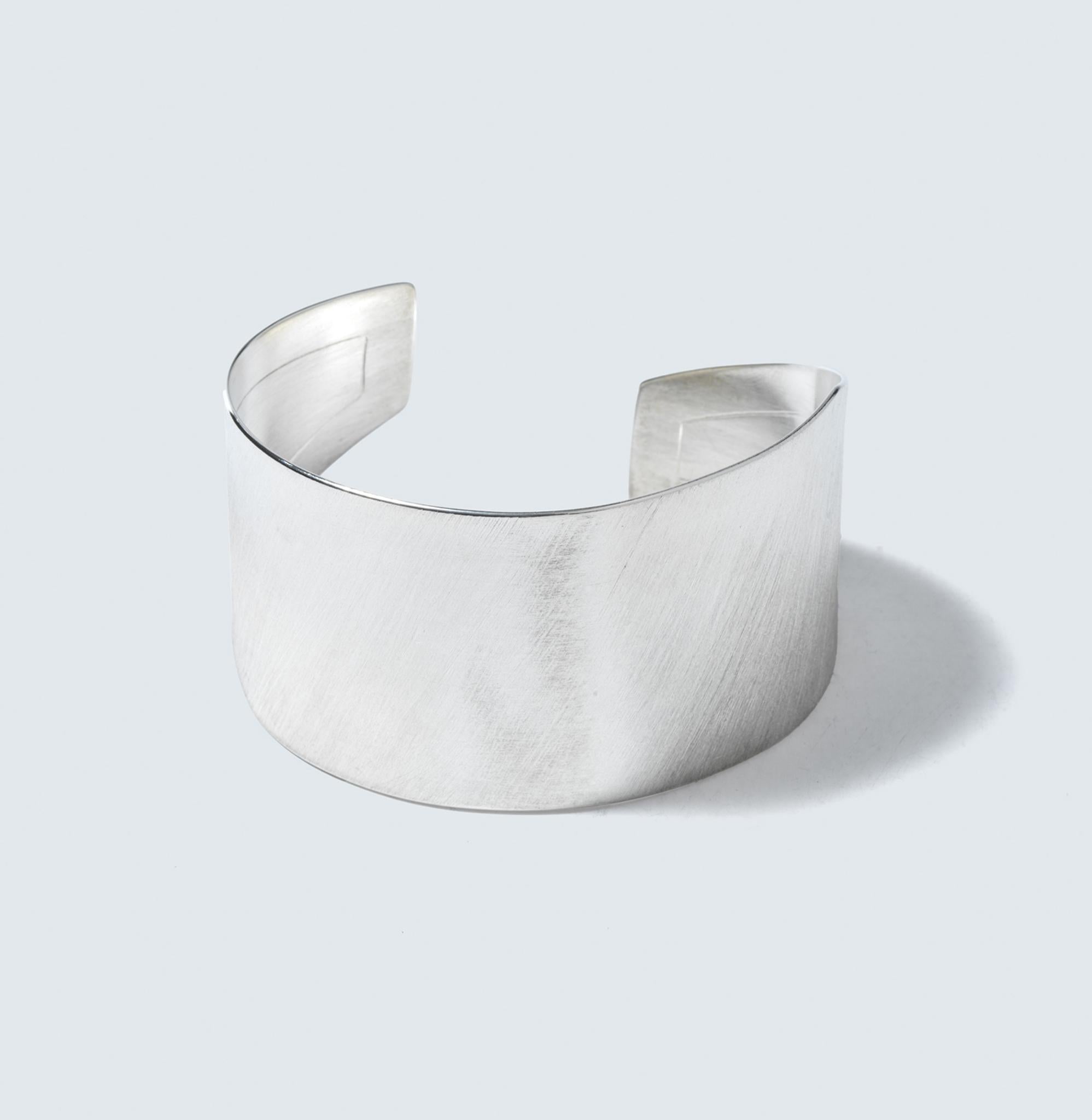 This is a sleek sterling silver bracelet with a broad, curved band. The surface of the bracelet is brushed, giving it a textured appearance that softly diffuses light. It has a satin-like luster that subtly catches the eye without being overly