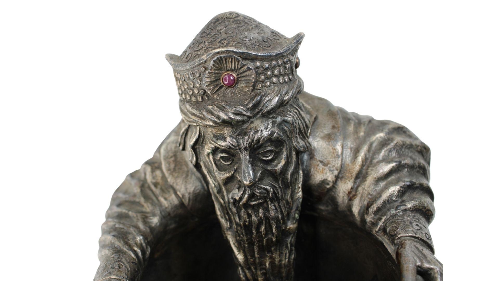 This stunning piece created by Pavel Ovchinnikov shows his amazing work in silver engraving.
This piece represents an old monk sitting while his beard falls into a cauldron.
On the monk's head is a hat with different shapes,  in the center of it