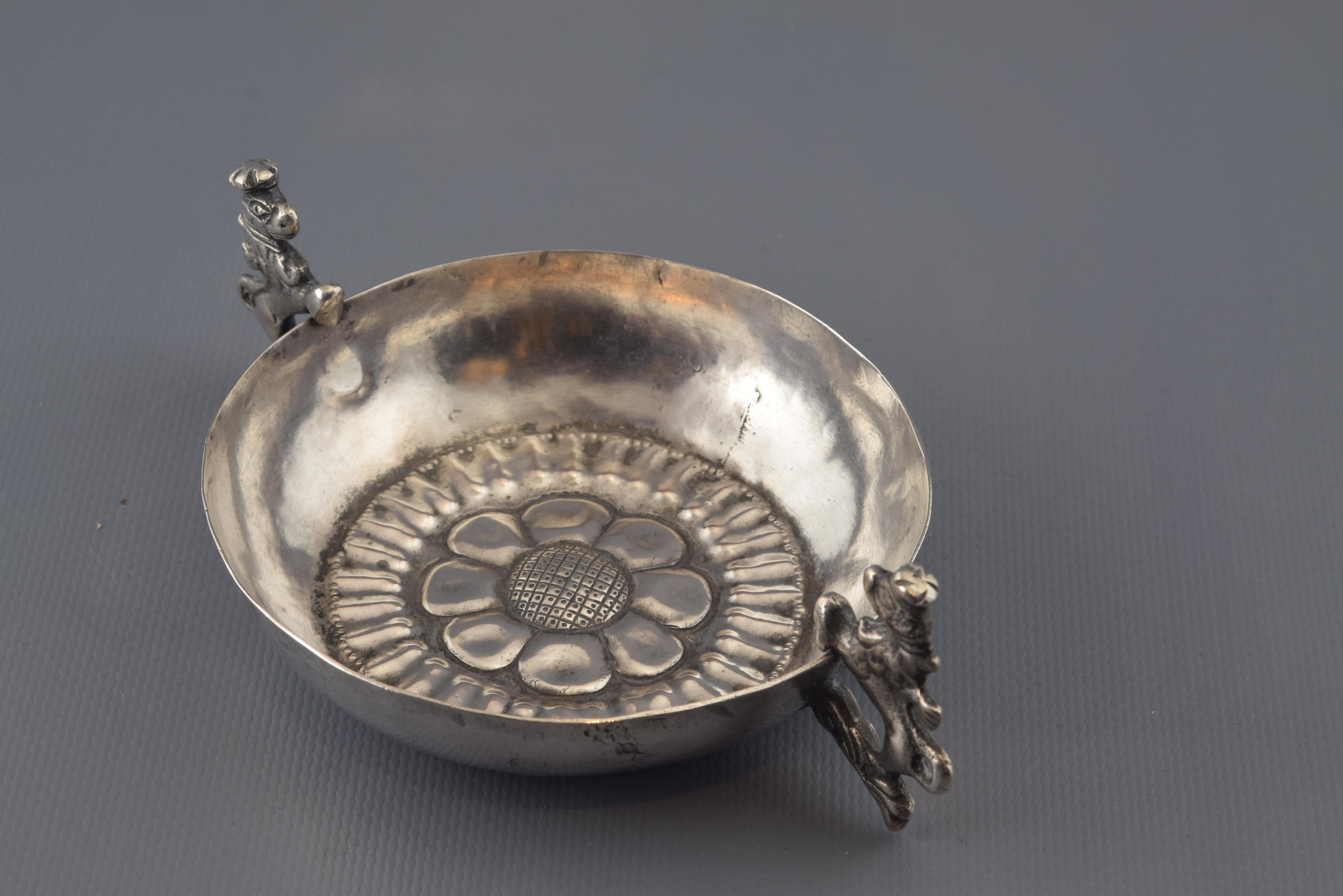 Sans signes distinctifs.
Gobelet en argent aux bords lisses, aux anses en forme d'animal couronné (lions rampants avec queue recourbée vers l'arrière) et à un décor au bas de la pièce de lignes encadrant une simple fleur. Dans l'argenterie