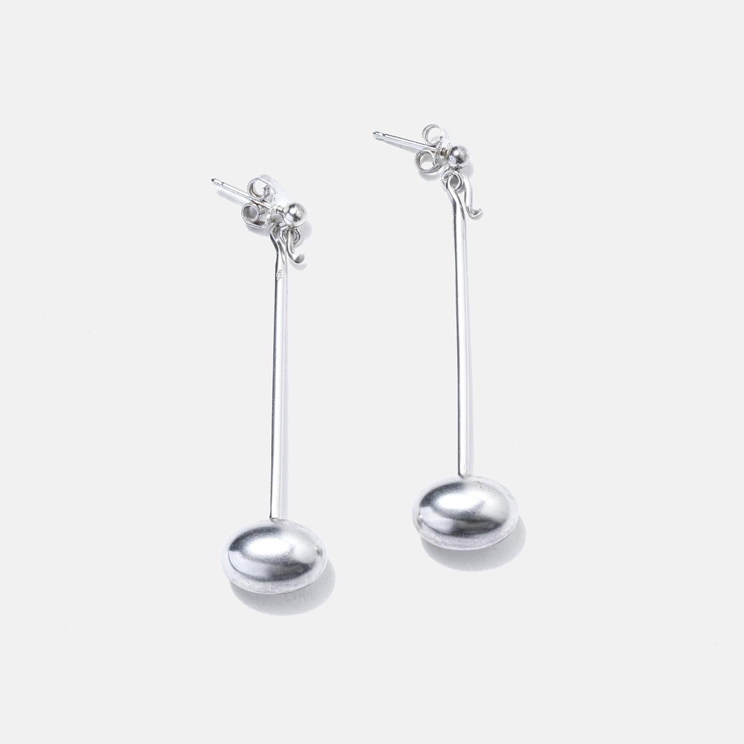 These earrings are crafted from sterling silver, featuring a sleek and minimalistic design. Each earring consists of a stud at the lobe from which a slender, straight rod extends, ending in a rounded, elongated bead that adds a touch of