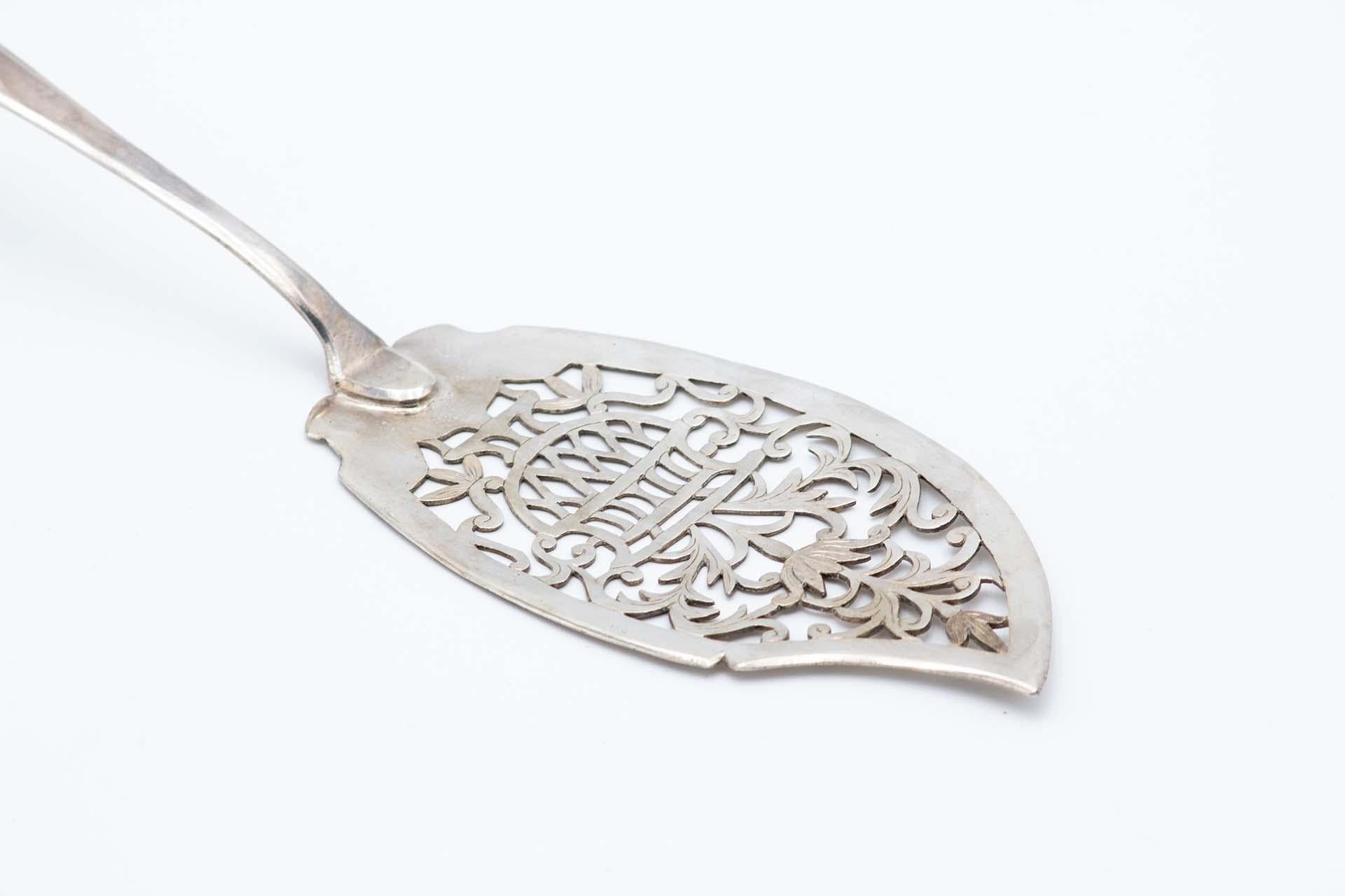 Description: Silver dessert spatula with floral motifs cut into the blade and a crimped cane-style handle. Metal: Silver. Origin: Portuguese. Brand: Porto Coroa. Weight: 140.89 grams. Dimensions: 31.4cmx4.6cm. Content: 833/1000. Assay Mark: Yes,