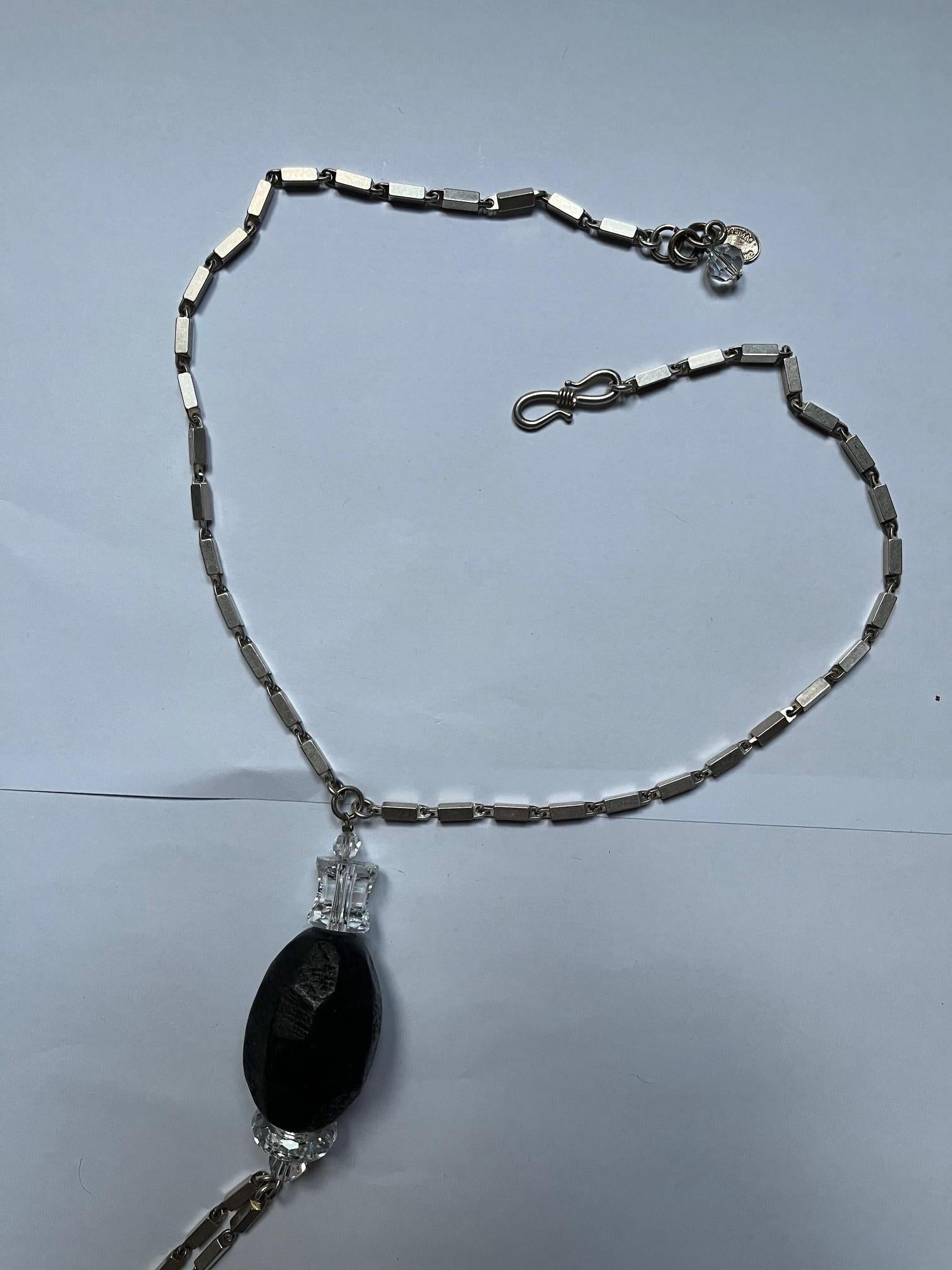 This beautiful lariat-style silver-toned necklace is a creation of Daniel Swarovski. The design is simple: a silver-toned square metal chain with 3 big dark-toned wood beads. Every bead is surrounded by typical clear Swarovski crystals. The 2 drop