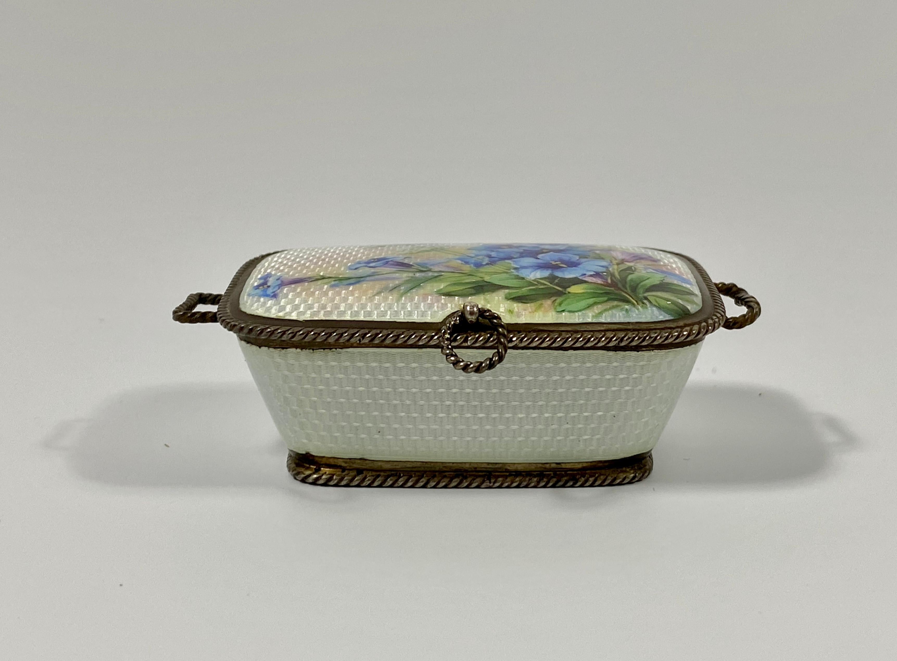 Continental silver and enamel box, date marks for 1911. The silver basket shaped box, engine turned, and painted to the cover with a spray of blue flowers. Having simulated rope twist silver handles, attached to the silver rope twist rims.
The