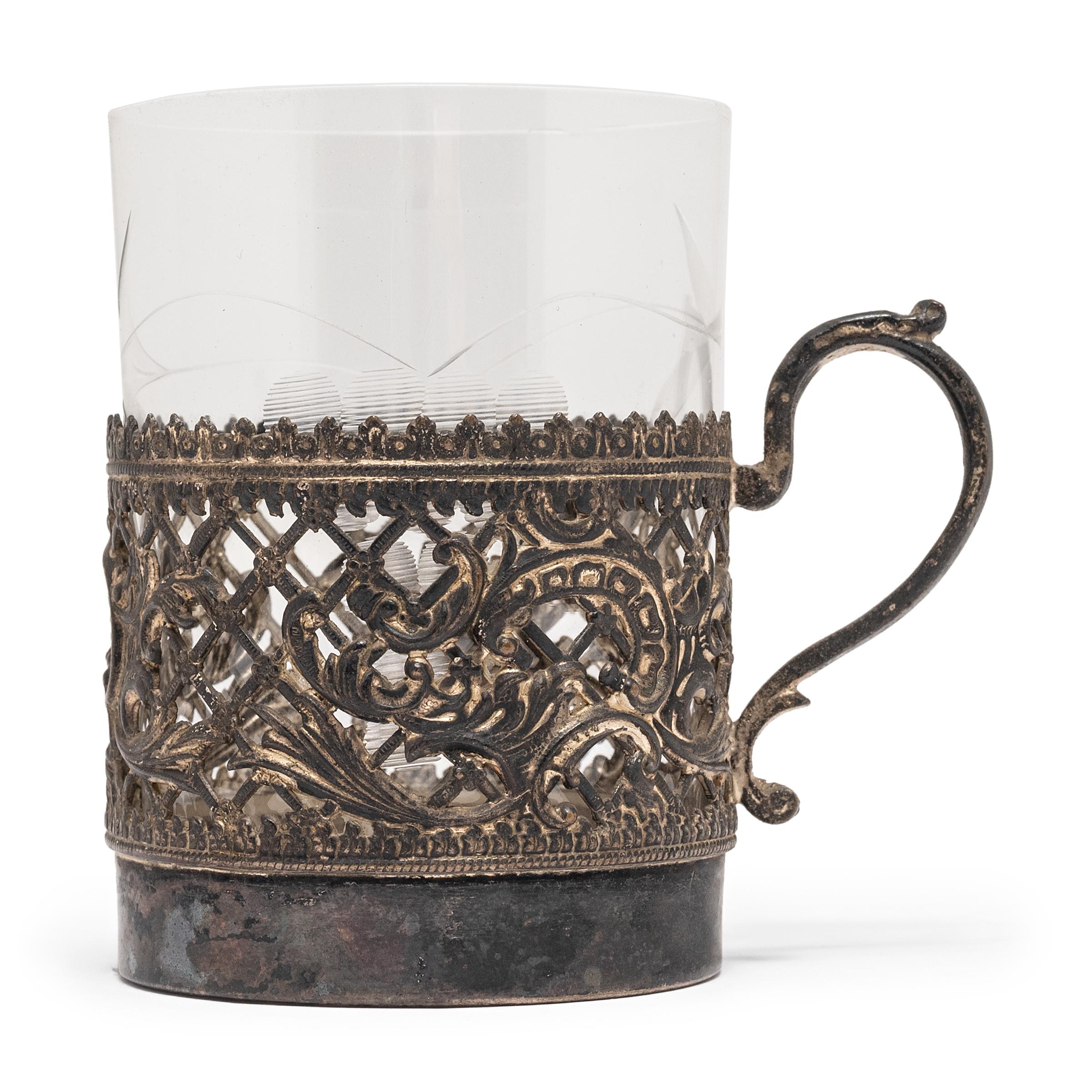 This 20th century European silver cup holder, set with a glass tumbler, boasts an exquisitely detailed filigree mount and scrolled handle. Relics of ancient filigree design have been found in Mesopotamia, dating the pressed metal technique as far