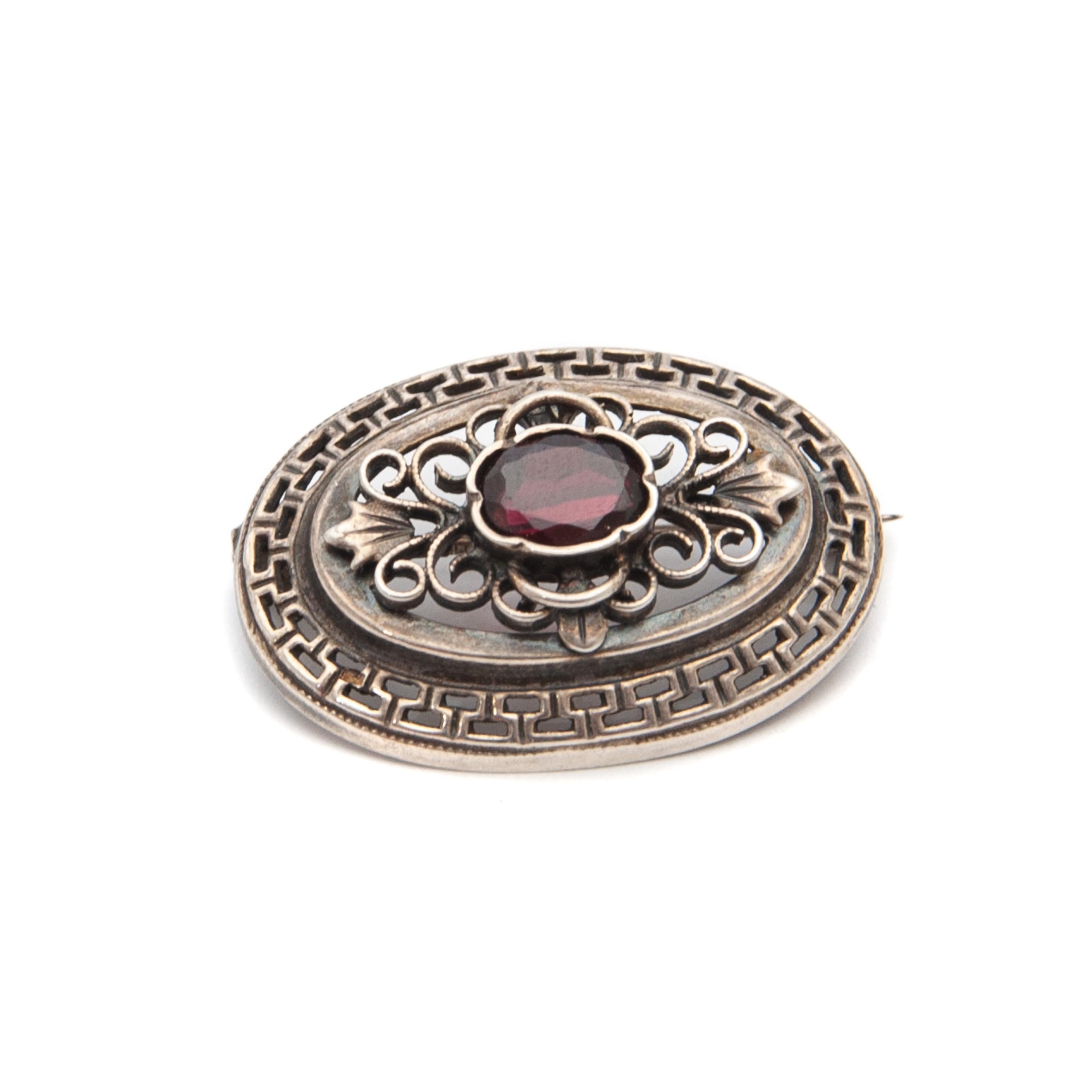This lovely mid-century openwork oval-shaped brooch is set with a faceted garnet in the middle. The silver has an openwork floral structure with swirling motifs, while the border has a repeating old Greek design. Which makes this romantic brooch