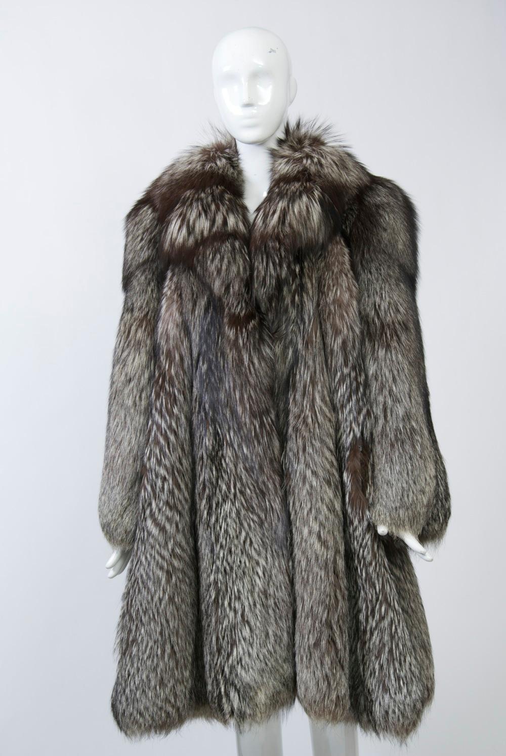 Knee-length silver fox coat featuring luxurious, full pelts, a notched collar and wide sleeves. The hem is scalloped following the natural curve of the vertical skins. Slash pockets. Two secure fur hook closure. Black and white print lining labeled