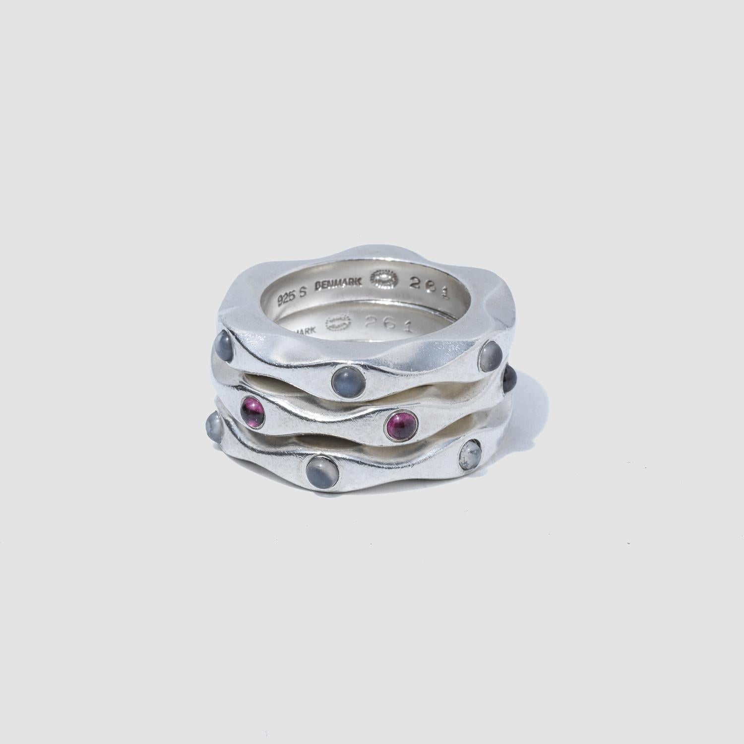 These sterling silver rings present a contemporary take on classic style, with one featuring three cabochon-cut garnets and the other two adorned with three moonstones, each gem set elegantly within the fluid design. Their wavy, contoured shape