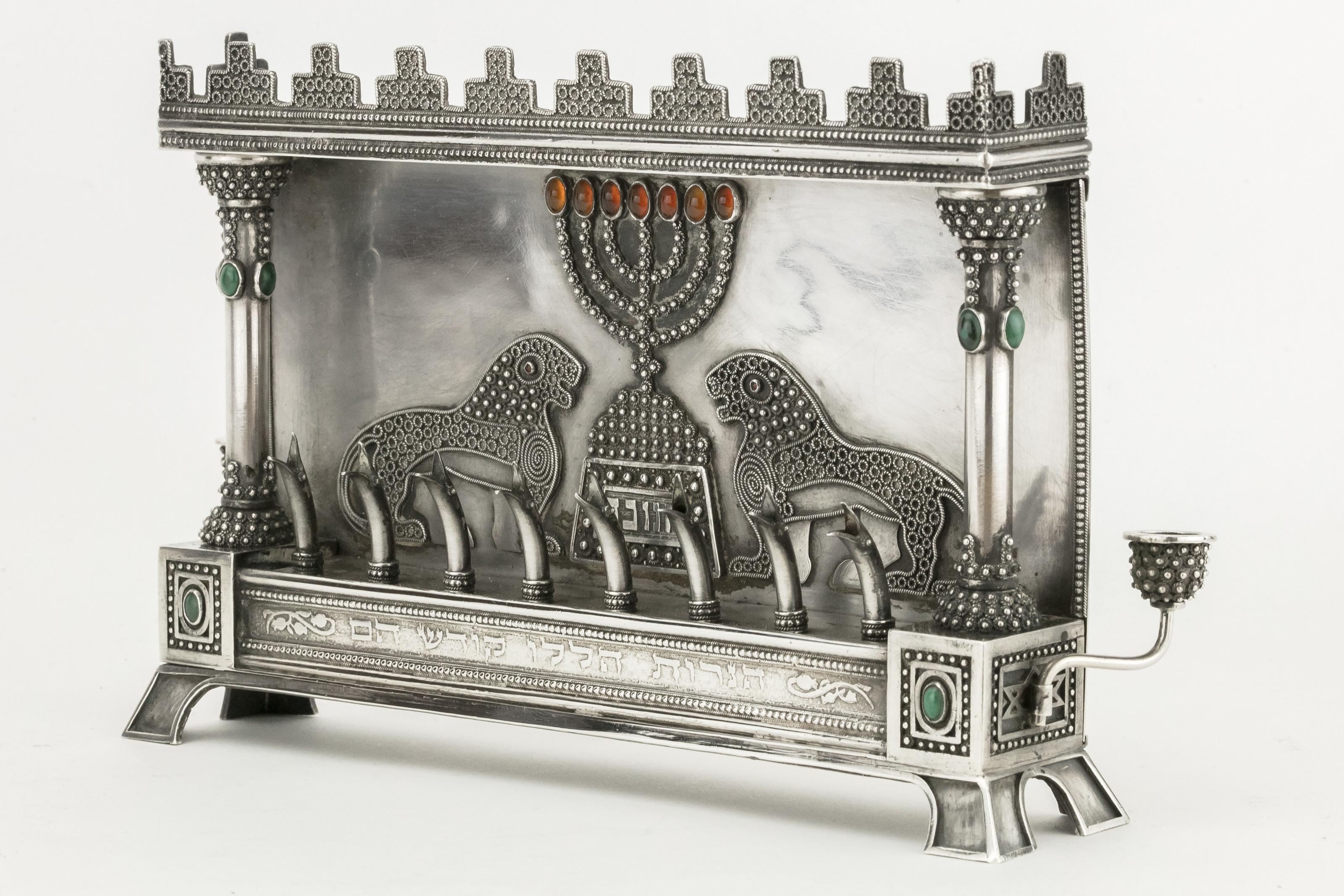 Silver and hardstone Hanukkah Lamp, Bezalel, Jerusalem, circa 1920.
The backplate is decorated with a pair of filigree lions flanking a Menorah with carnelian cabochon flames, under a pediment and between columns mounted with malachite, the front
