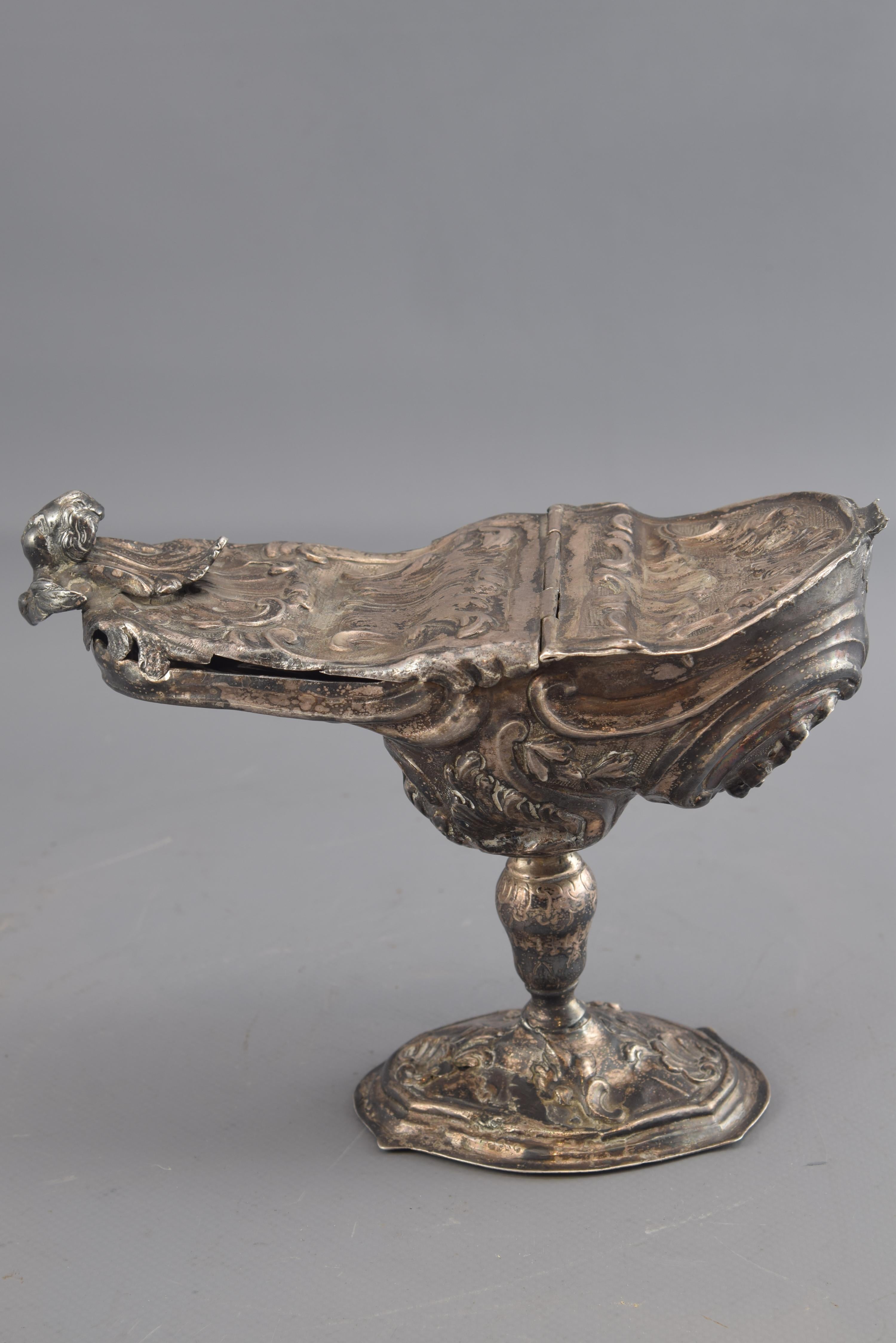 Naveta. Silver. Possibly Portugal, 18th century.
Base with counterweight. With hallmarks.
Naveta with oval foot decorated with relief elements and moldings, balustrade foot and body with light boat shape, presenting a hinged lid in the front, a